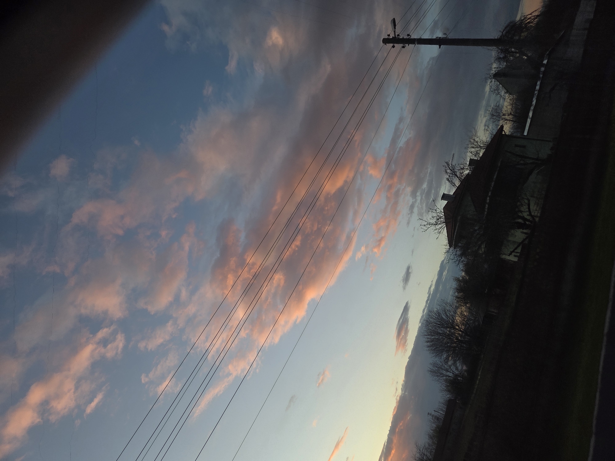 The image is a low-angle shot of a scene in Koilovtsi, Bulgaria. The focus is on a twilight sky with pink-tinged clouds. In the foreground, power lines stretch across the frame. To the right, a silhouette of a house with a chimney and bare trees is visible, along with a telephone pole. A dark, undefined shape occupies the left corner of the frame, potentially a part of a structure or object the image was taken from. The overall lighting is low, suggesting dusk or dawn.