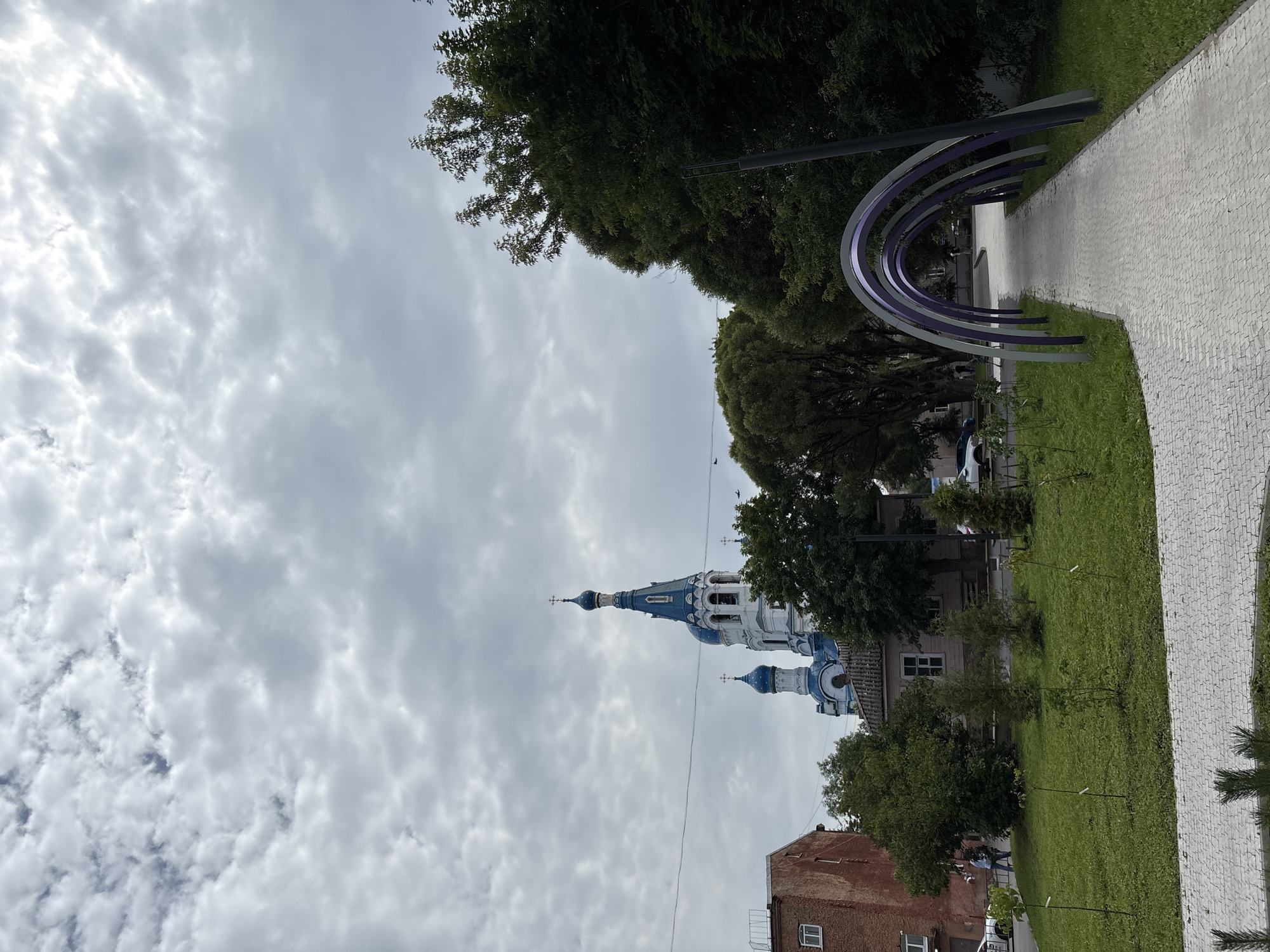 This is an exterior, eye-level, vertical shot of a public walkway with an arched entrance in Gatchina, Russia. The walkway is paved with light-colored interlocking bricks and is lined on both sides with green grass and young trees. A series of five purple and grey arches form an entrance to the walkway. On the left side of the walkway, there is a grassy area with several small trees. Behind this area, a red brick building stands with a white-framed window. Further back, a white church with blue domes and crosses is visible. Trees and more buildings are present in the background, and the sky is overcast with grey clouds. Two people are walking on the path in the distance.