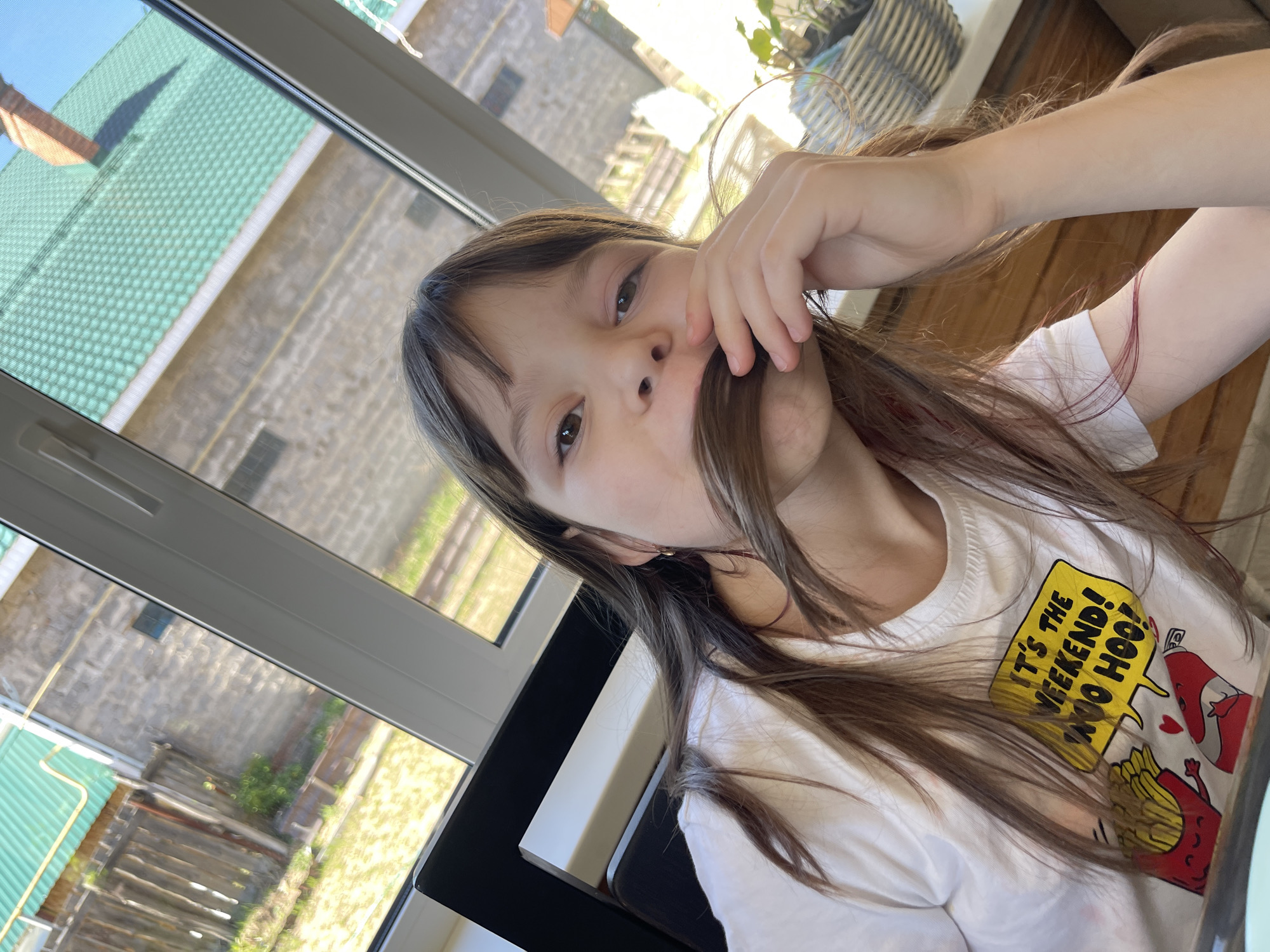 A young girl with long brown hair holds a strand of her hair over her upper lip, creating a makeshift mustache. She wears a white T-shirt with a graphic that reads, "IT'S THE WEEKEND! WOO HOO!". She is indoors, near a window that provides a view of buildings with green roofs, indicating a residential area. Based on location metadata, this image was taken in the city of Chaikovsky, Russia.
