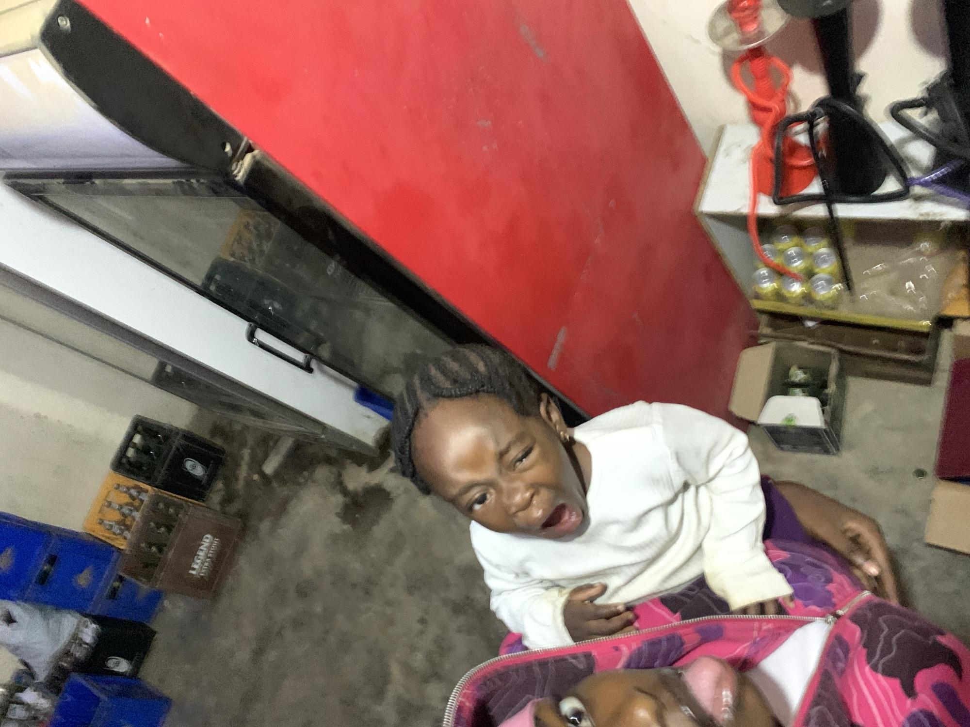 A canted, high-angle shot shows two young children in an indoor setting, apparently in the city of New Stadium, Nigeria. The child in the foreground is partially visible, displaying an open eye. The other child is looking upwards, their mouth open in an expression. They are wearing a white, long-sleeved shirt and are seated on a surface covered with a pink and purple patterned fabric.
In the background, a stack of blue, yellow and brown crates is visible, along with a white refrigerator, and a red-colored counter or table. A white table with cans stacked on top sits to the right, accompanied by a lamp. The floor is concrete.