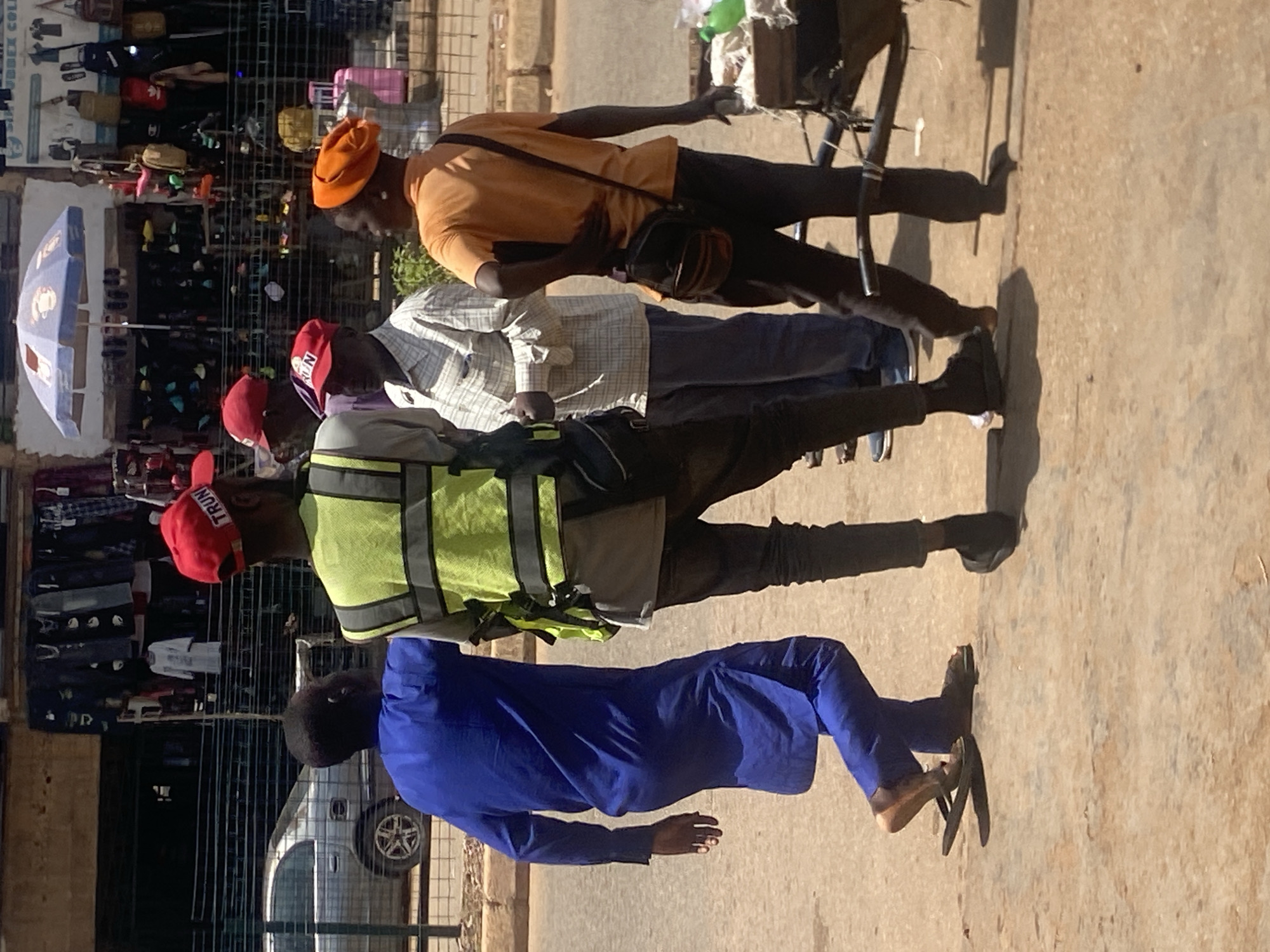 In Bukuru, Nigeria, four individuals are depicted standing on a dirt and asphalt road in front of a market stall with clothing displayed. A silver sedan is visible parked behind a metal fence in the background. The individual on the far left wears a vibrant blue garment and dark sandals. To their right, a person in a bright yellow reflective vest over a light-colored shirt and dark pants is positioned, wearing a red cap with "TRUMP" printed on it. Next to them, a man in a plaid shirt and dark pants stands. The individual on the far right wears an orange beanie, an orange t-shirt, and dark pants, carrying a dark shoulder bag. A cart with items on it is positioned to their right.