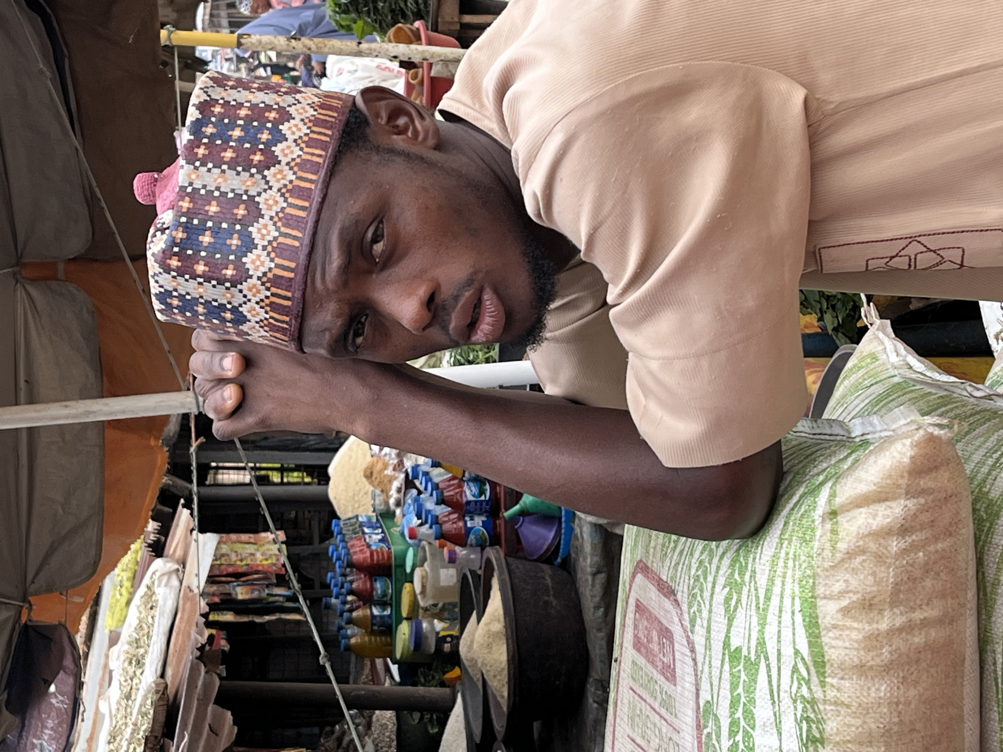 The image depicts a man wearing a traditional cap and a light brown robe, leaning on a stack of large rice bags at an outdoor market. His hands are clasped in front of him. Behind him, various market stalls are visible, displaying bottled beverages, sacks of grains, and other goods. The setting appears to be a bustling marketplace. The man's gaze is directed towards the camera. The overall atmosphere suggests a vibrant commercial environment.