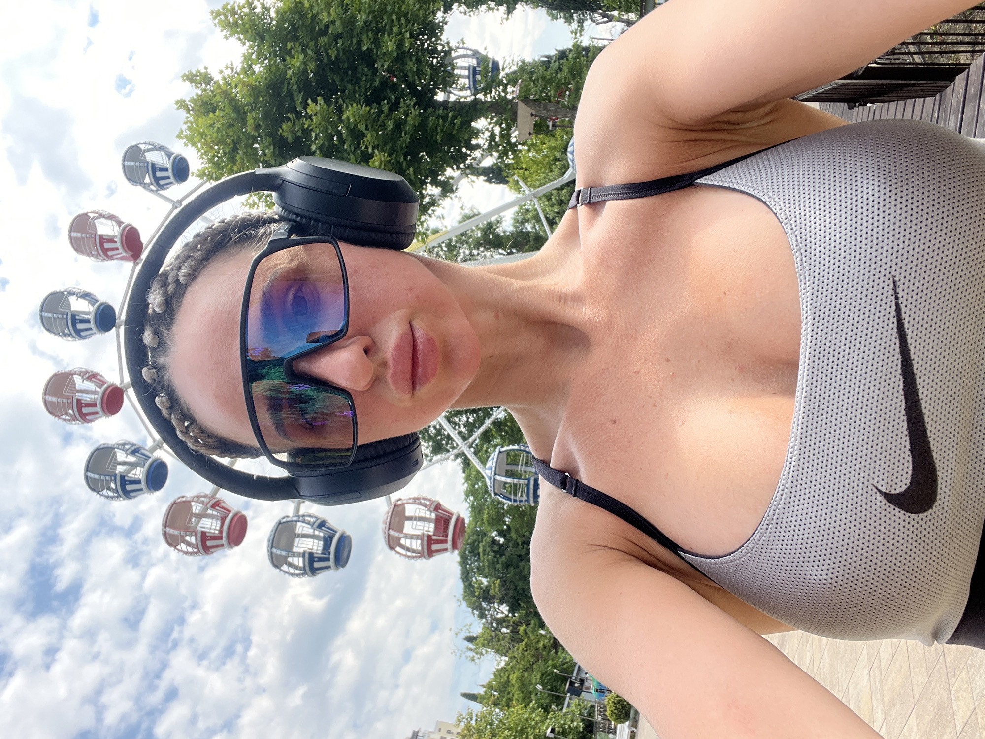This is a selfie taken by a woman in Sochi, Russia. She is wearing a grey Nike sports bra and black headphones. She has on large, reflective sunglasses that obscure her eyes. Her hair is styled in braids. In the background, a Ferris wheel is visible, with some of its colorful gondolas in view. Lush green trees and a bright, partly cloudy sky suggest it is daytime and the weather is pleasant. The overall impression is of someone enjoying an outdoor activity, possibly at an amusement park or entertainment venue. The composition of the photo, with the subject close to the camera and the background elements blurred, emphasizes the individual while still providing context of her surroundings.