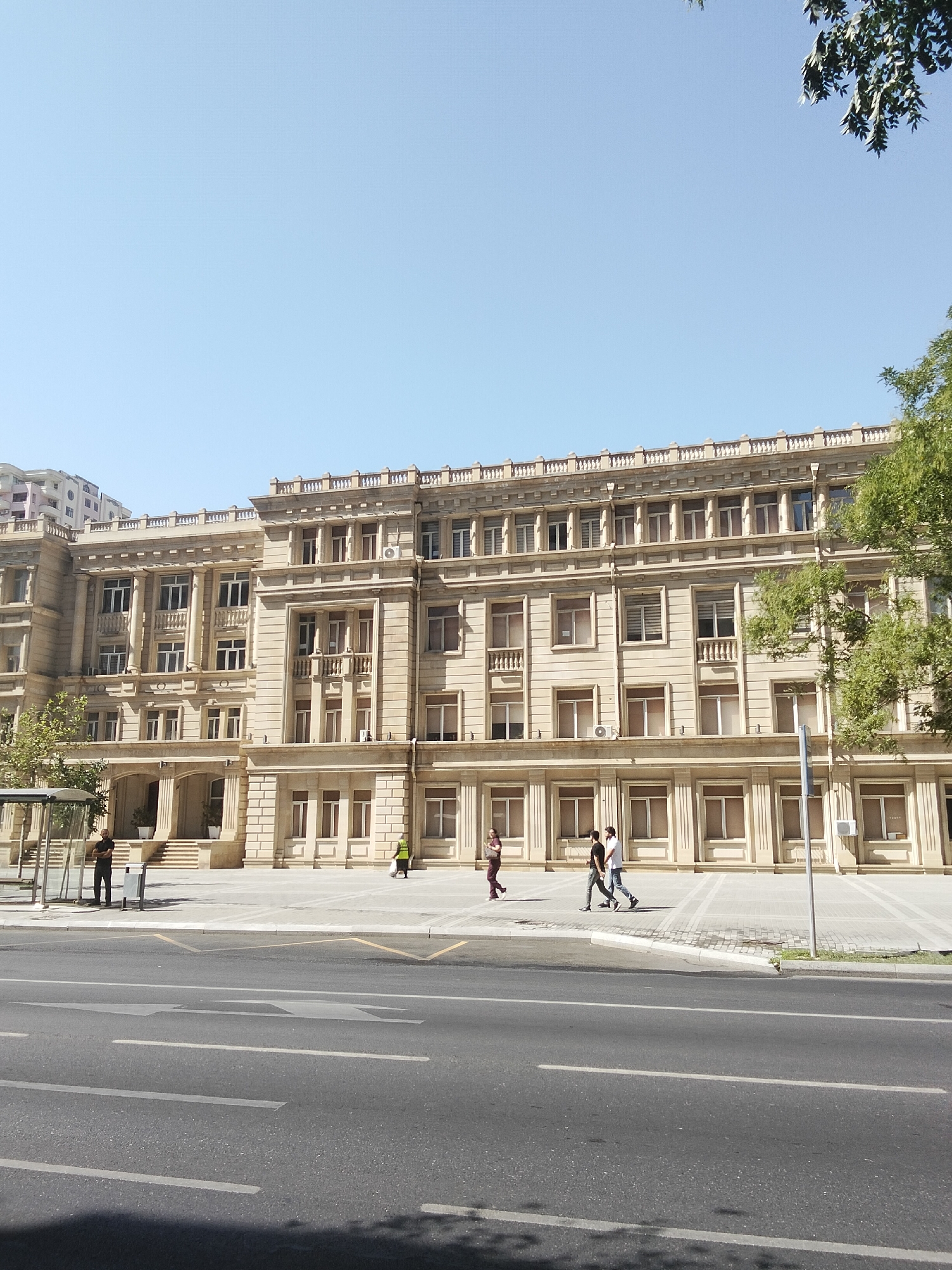 An exterior, eye-level, full shot captures a section of a grand, multi-story sandstone building in Baku, Azerbaijan, likely an administrative or governmental structure. The building features classical architectural elements, including ornate cornices, columns, and repetitive window patterns on multiple floors. In the foreground, a wide, asphalt road runs horizontally across the frame, marked with white and yellow lane lines and directional arrows. To the left, a bus stop shelter with a glass facade is visible, and a lone individual stands near it. Further to the right, along the sidewalk in front of the building, three individuals are walking, two of them in conversation. Another person in a high-visibility vest is seen carrying a bag near the building's entrance. Lush green trees flank the right side of the building, their leaves catching the sunlight. The sky above is a clear, pale blue, indicating a sunny day. The overall impression is one of a well-maintained urban environment, showcasing a blend of historic architecture and modern infrastructure.