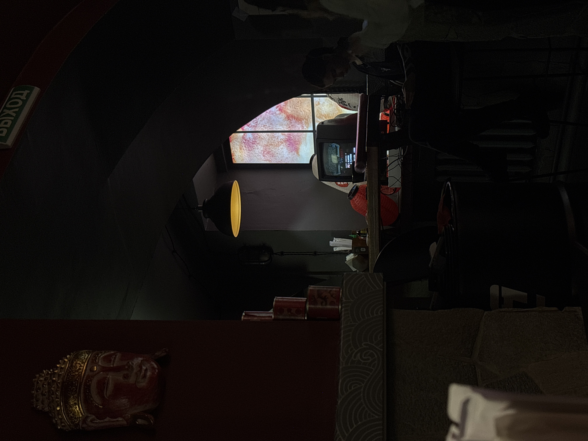 The image shows an interior view of a dimly lit space, seemingly a bar or lounge in Perm, Russia. The space features an arched entryway with a screen displaying a soft, abstract cloud-like pattern. A person sits at a desk with a CRT monitor. Cans or small containers are stacked on a surface. A Buddha head statue is displayed on a surface in the lower portion of the scene. An exit sign with text in Russian is visible in the upper left corner. Furniture includes tables and chairs.