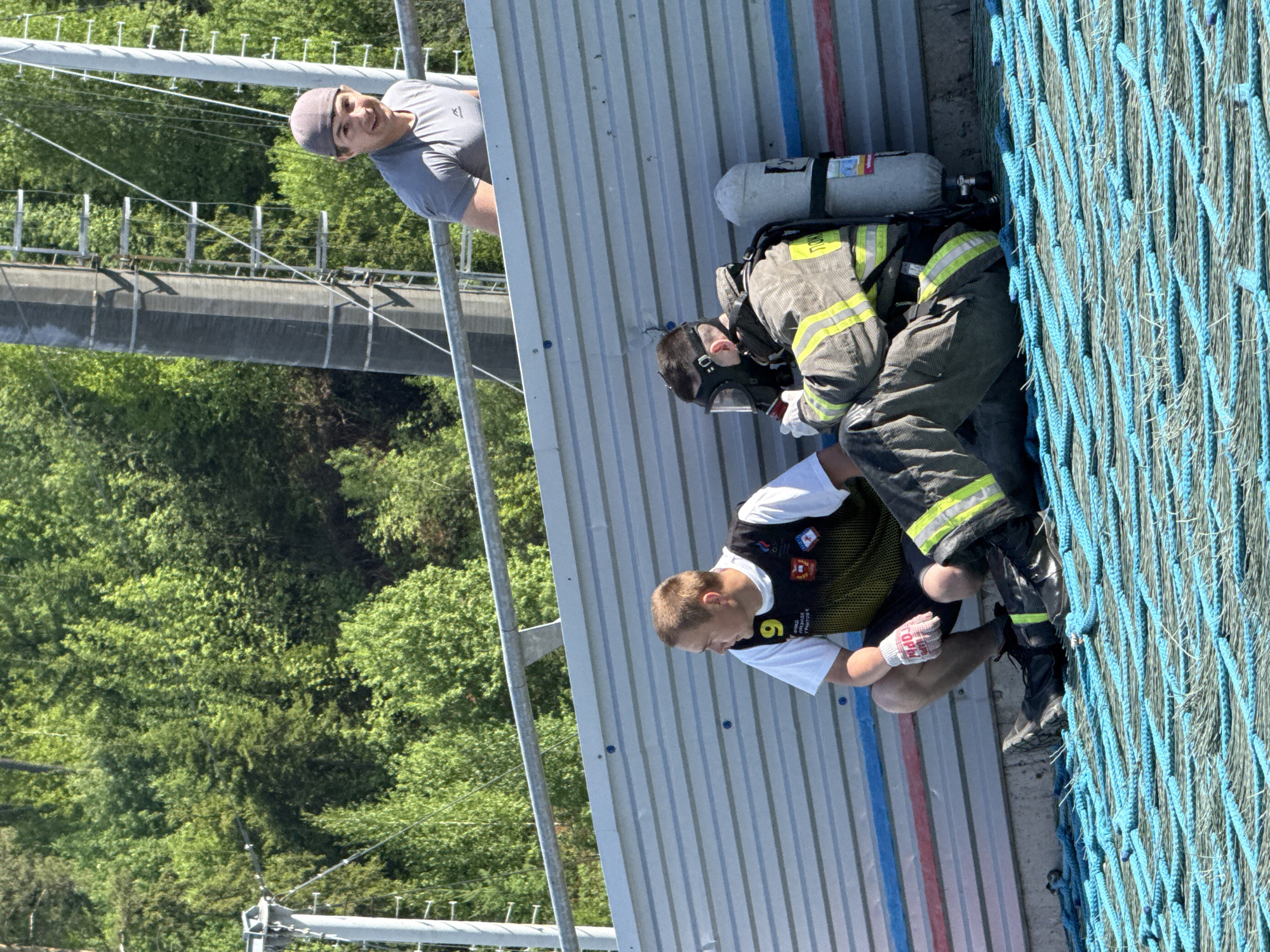 The image captures three individuals in an outdoor setting, likely in the city of Unknown, Unknown. A man with a gray hat and gray t-shirt stands at the top left, possibly observing. Below him, a firefighter in full gear, including an oxygen tank and mask, is kneeling. Another individual wearing a white t-shirt and a black vest with the number "9" on it is kneeling as well, close to the firefighter. The individuals are positioned against a gray corrugated metal wall with blue and red vertical stripes near the base, adjacent to a blue rope net. The backdrop features trees and a tall structure in the distance.
