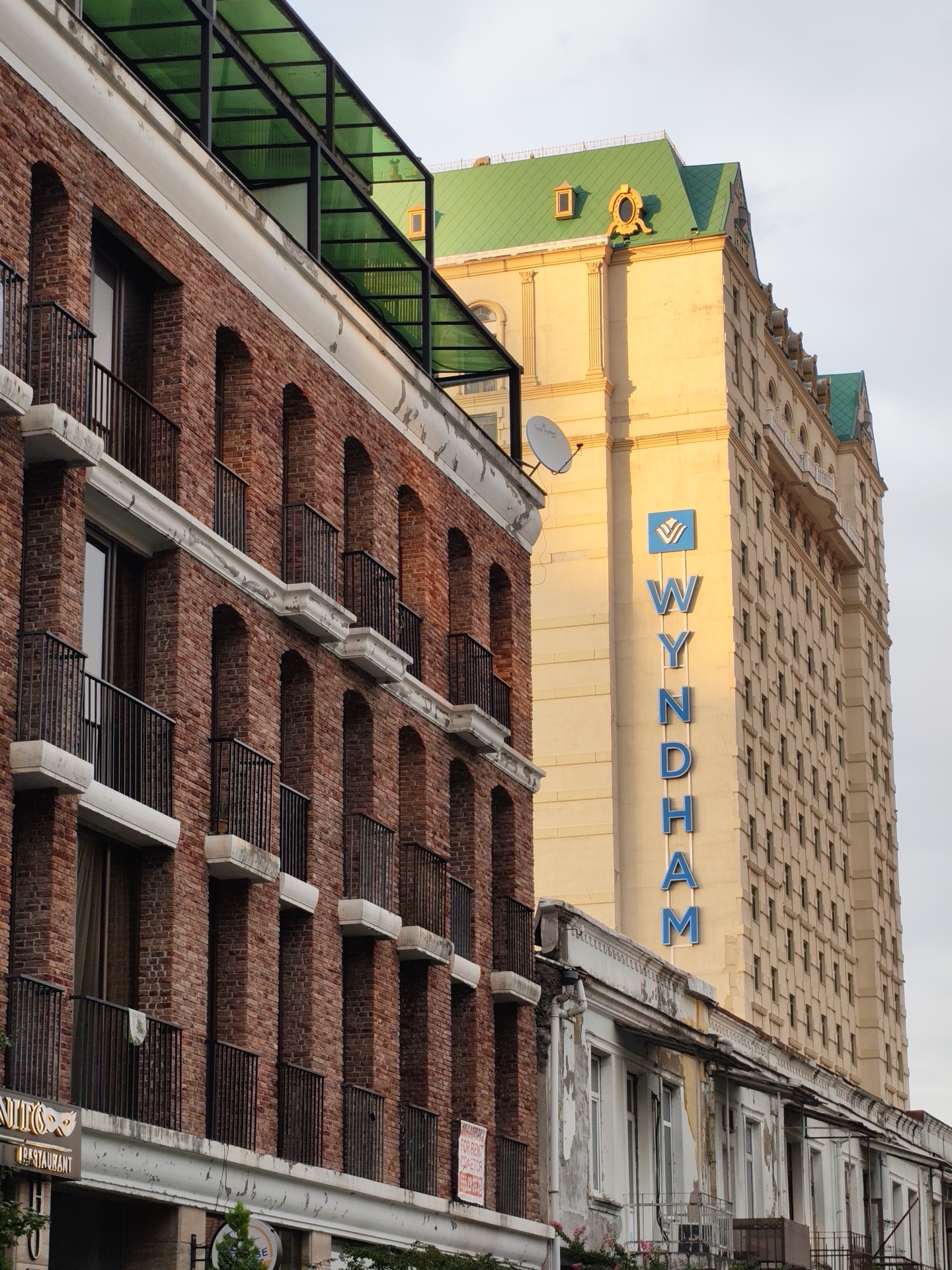The image shows a street view in Batumi, Georgia, with two buildings prominently featured. On the left is a red brick building with arched windows and balconies, topped with a green, translucent awning. To the right, a tall, light-yellow building with a green roof rises, displaying a large vertical "WYNDHAM" sign in blue letters. The sky is overcast. In the foreground, a smaller, older building with peeling paint and balconies sits in front of the Wyndham. A satellite dish is visible on the older building. Signs for "RESTAURANT" and "FOR RENT" are also visible on the lower parts of the buildings.