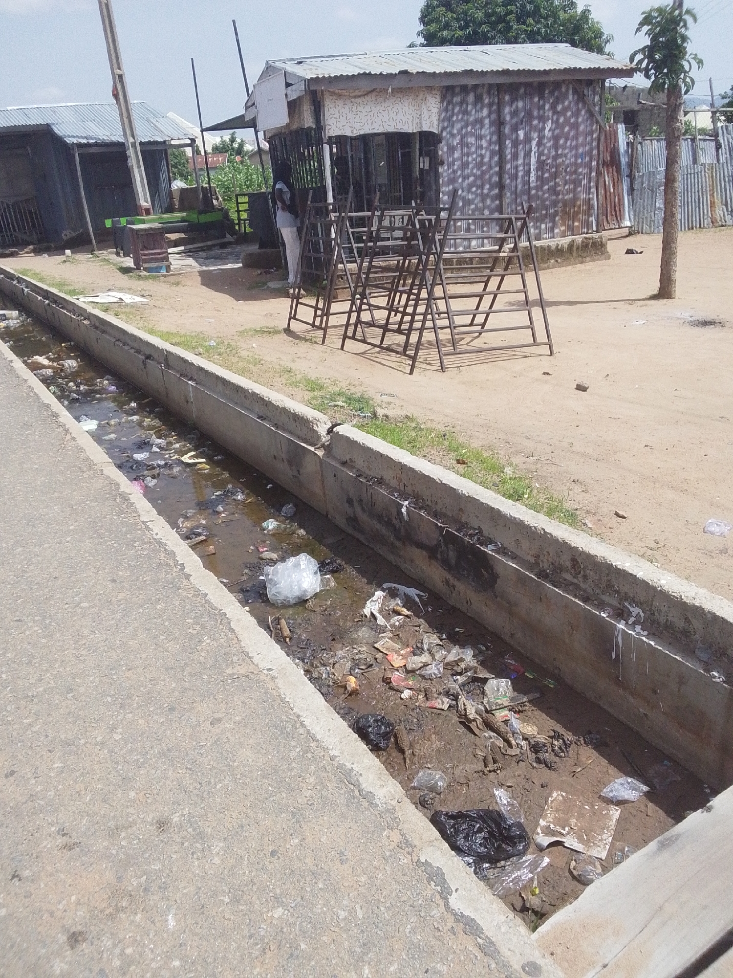 The image depicts a drainage ditch filled with water and a significant amount of refuse, including plastic bags, paper, and other debris. The ditch runs alongside a paved area and a dirt path. In the background, there are several structures that appear to be small shops or market stalls constructed with corrugated metal and wood. A person wearing light-colored clothing stands near one of the stalls. In front of the stalls are several metal frames, possibly for display or seating. The environment is dusty and dry, with some sparse vegetation and trees visible. The overall scene suggests a location within a city, potentially in an area with less developed infrastructure, consistent with parts of Jalingo, Nigeria, where such open drainage and waste management issues can be observed.
