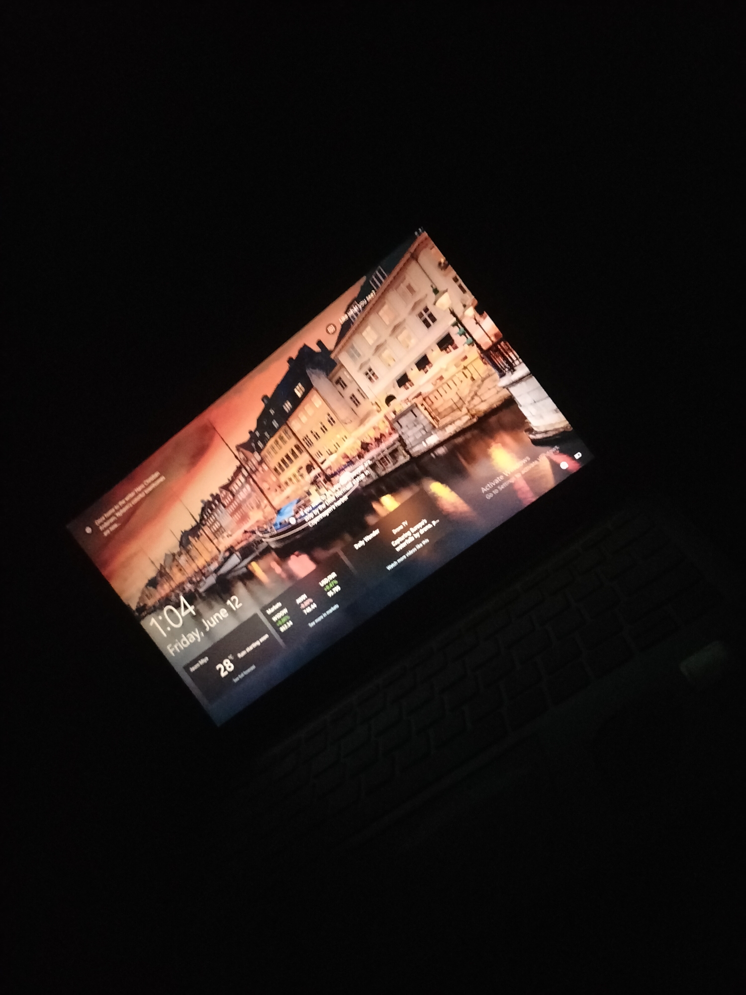 A laptop screen displays a lock screen with a scenic view of a European city at sunset, featuring buildings and a canal. The time is 1:04 AM on Friday, June 12. Below the time, a weather icon indicates 28°C with "Rain starting soon." A section to the right shows stock market tickers for USD/INR, MSFT, and AMZN, with their respective percentage changes. Another section displays news headlines and the option to "Watch more videos like this." The words "Activate Windows" are faintly visible in the bottom right corner. The laptop is angled and placed on a black keyboard. The image is dark, with only the laptop screen illuminated. There is no visible presence of Jalingo, Nigeria in the image.
