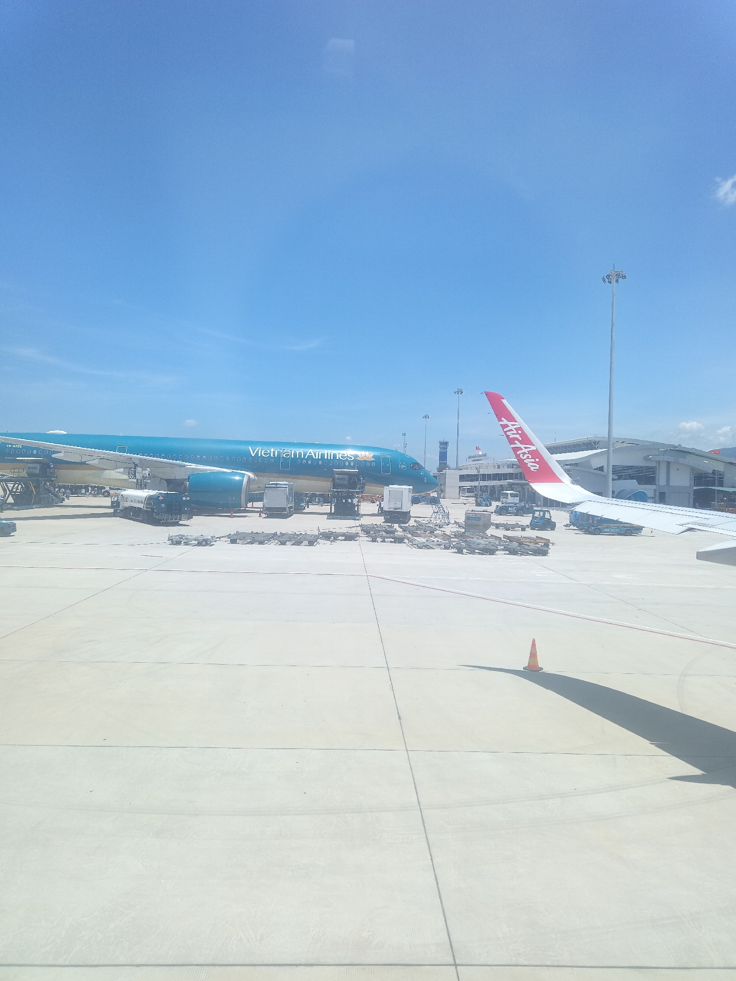 The image depicts an airport tarmac under a clear blue sky. A large, turquoise airplane with "Vietnam Airlines" written on its side is parked. Adjacent to it, a wing of another airplane, bearing the "AirAsia" logo in red and white, extends into the frame. Several service vehicles, including a fuel truck and various carts, are positioned around the Vietnam Airlines aircraft, suggesting it is undergoing servicing. In the background, an airport terminal building and a tall lighting mast are visible. The tarmac itself is paved with concrete, marked with lines, and a solitary orange traffic cone casts a long shadow. The scene is static, capturing a moment of activity at the airport. The location is identified as Bắc Cam Ranh, Vietnam.