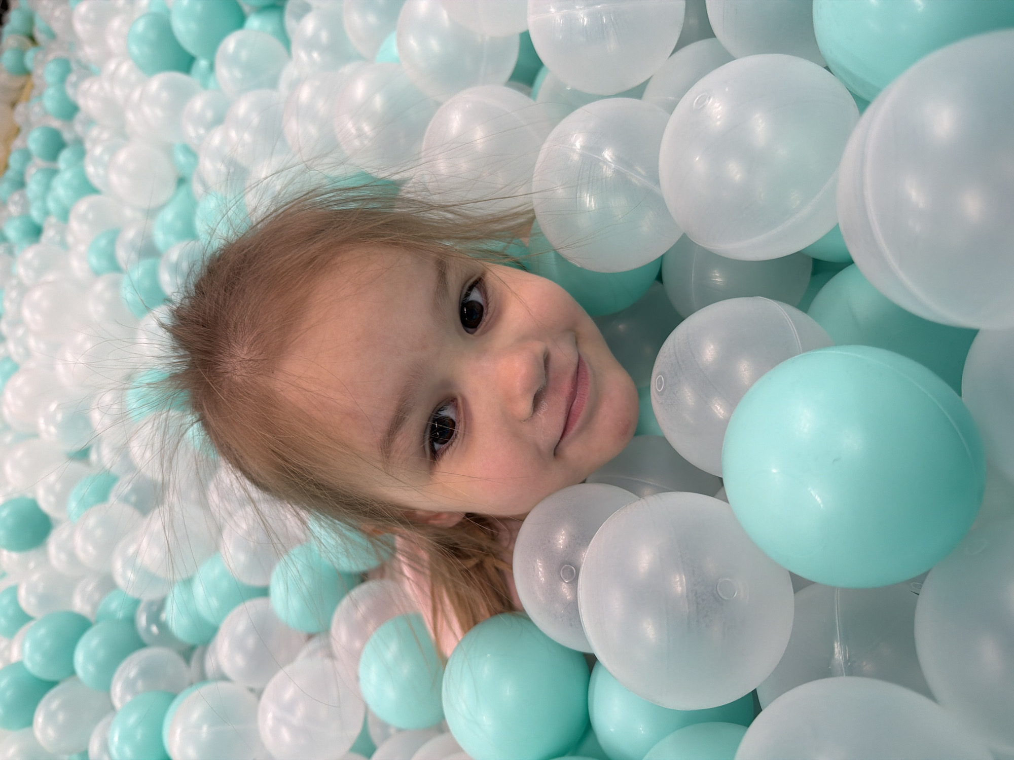 A young girl is surrounded by a ball pit of white and light blue balls. The girl has light brown hair and is smiling. Her head is tilted to the side. The balls are translucent. The photo appears to be taken close up. It is not possible to verify the location of the image based on the visual data.
