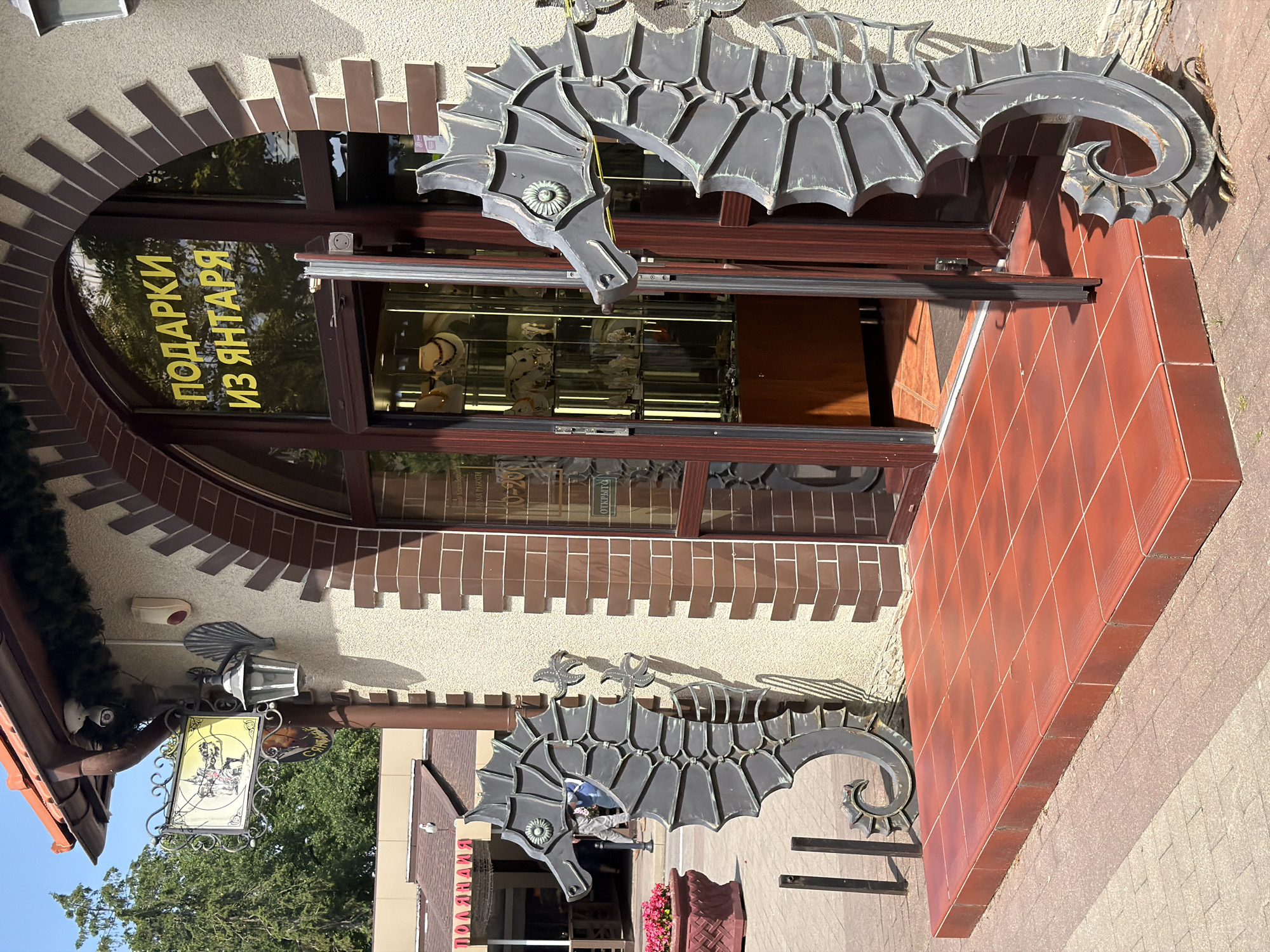 The image depicts the entrance to a shop in Svetlogorsk, Russia, as indicated by the Cyrillic text "ПОДАРКИ ИЗ ЯНТАРЯ" (Gifts from Amber) above the entrance. The shop appears to sell amber jewelry and souvenirs, with display cases visible inside. The storefront is decorated with two large, stylized metal seahorse sculptures flanking the entrance. The entrance itself is a double doorway with a glass facade, and the door is ajar. A red tiled platform leads up to the entrance. The building has a textured beige facade with brown brick accents around the arched doorway. To the left of the entrance, a sign with a historical or fantastical scene is mounted near a lamppost. The surrounding environment suggests an outdoor public space, possibly a street or promenade, with trees and other buildings visible in the background. The lighting indicates it is daytime with sunlight casting shadows. There are no people prominently featured in the foreground, though a few individuals are distantly visible inside the shop and in the background.