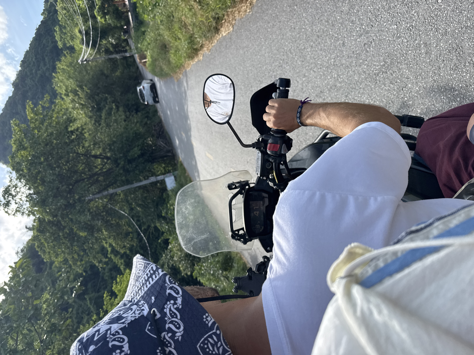 In Nam Hoa Lư Ward, Vietnam, a person wearing a white t-shirt and a blue bandana-patterned bucket hat rides a motorcycle on a paved road. The rider's left hand is visible on the handlebars, adorned with a woven bracelet. A motorcycle windshield is present, and a side mirror reflects the rider's face. The surrounding environment consists of lush green foliage and a mountainous landscape under a bright, partly cloudy sky. A white car is visible in the distance on the road.