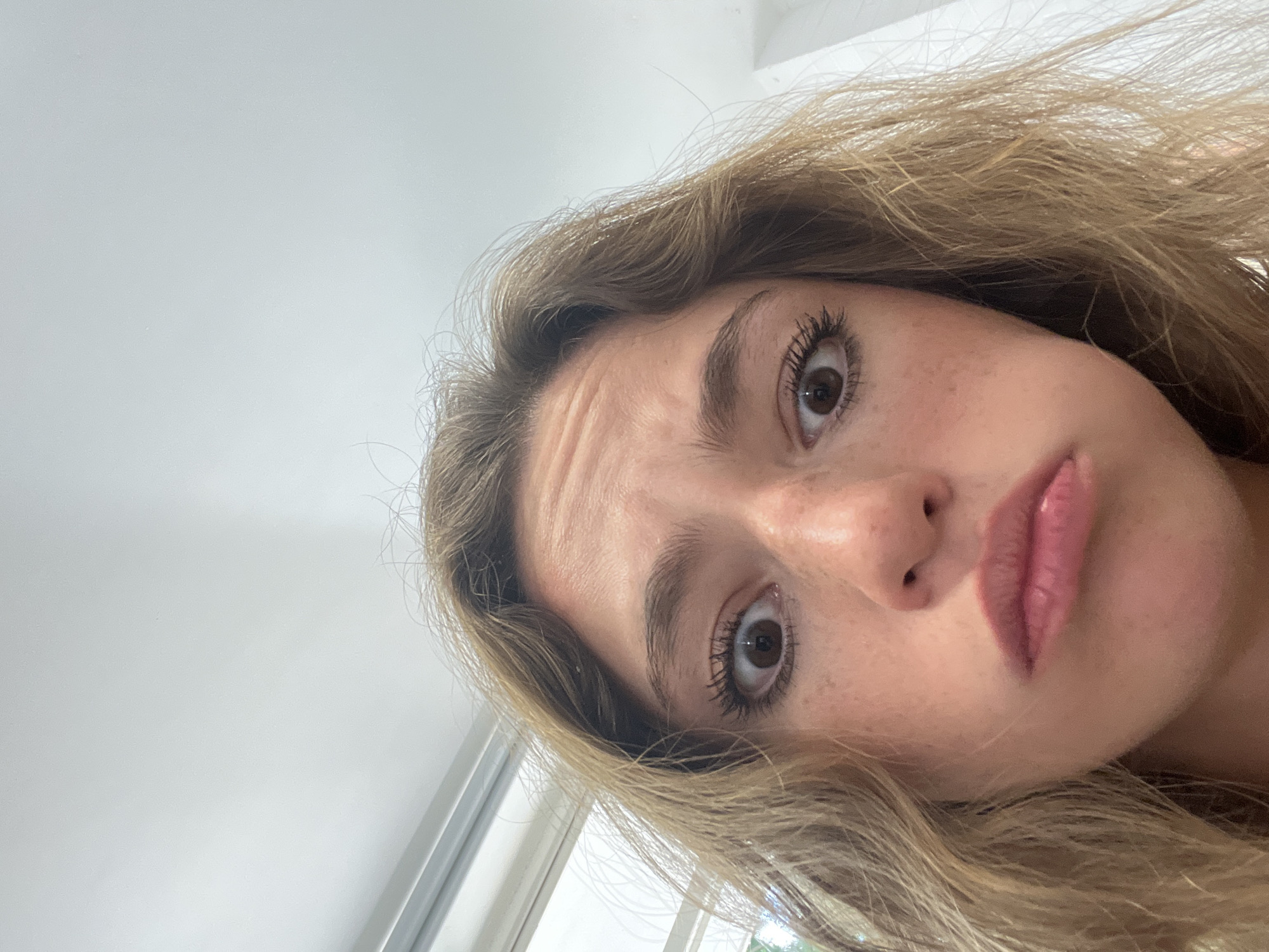 A close-up, slightly tilted image shows the face of a young woman with light brown hair and fair skin. Her expression appears quizzical, with furrowed brows and a slight pout. She has brown eyes, freckles on her nose and cheeks, and wears mascara and lip gloss. The background is primarily a plain white surface, with a portion of a window visible in the lower left corner. There is no indication of Puebla, Mexico, or any specific city location.