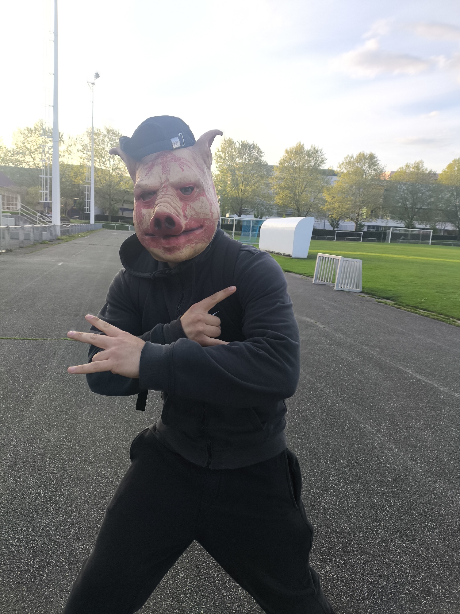 A person wearing a pig mask and a black baseball cap, hoodie, and sweatpants stands on an asphalt track, making hand gestures. The person is positioned in front of a green soccer field with soccer goals and a white sports shelter. In the background, there are trees and buildings under a partially cloudy sky. The location is Schiltigheim, France, as evidenced by the typical European architecture and sports facilities.