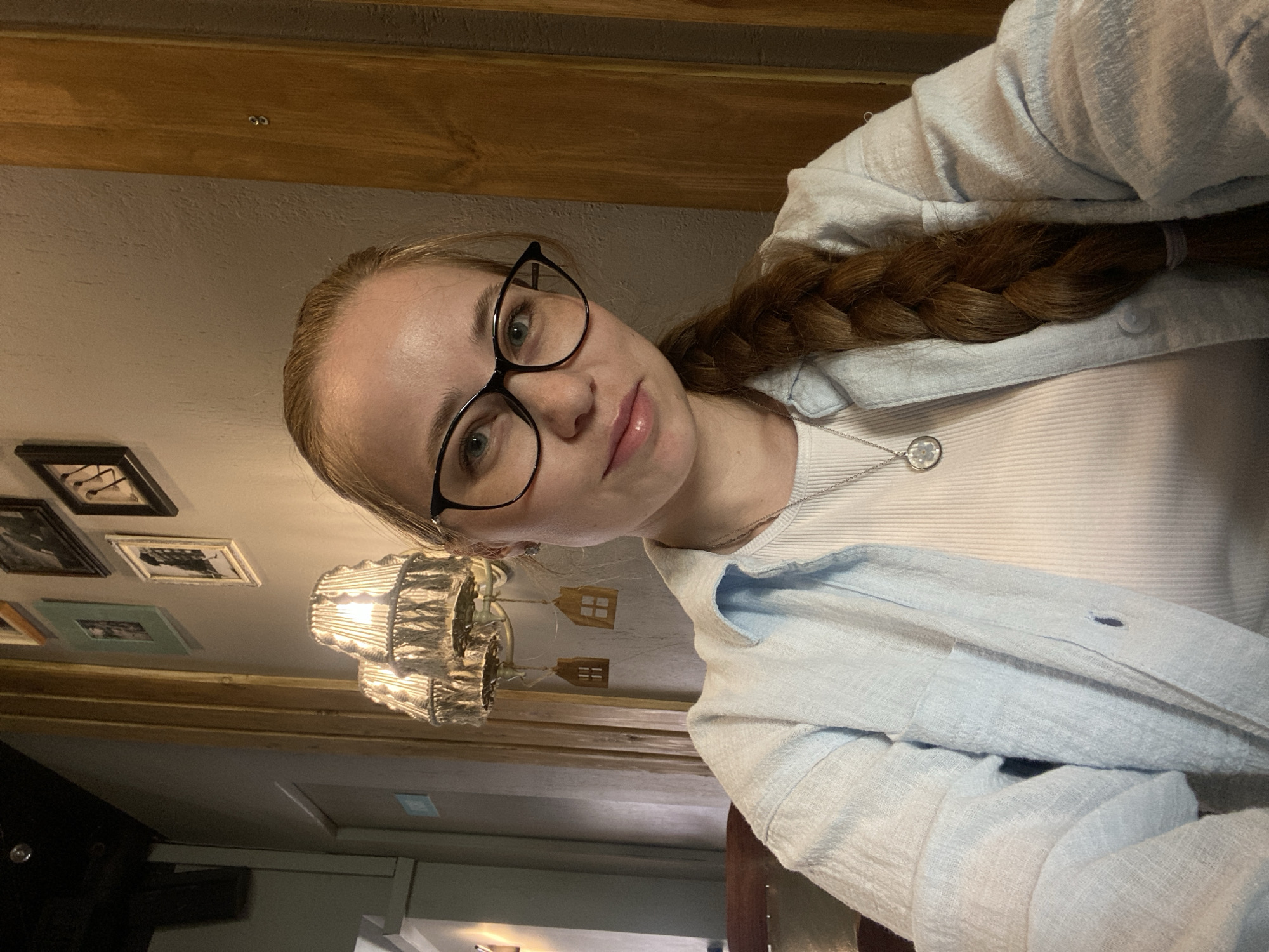 A young woman is captured in a close-up, tilted self-portrait, likely indoors in Saint Petersburg, Russia. She has fair skin, light-colored hair styled in a long braid over her right shoulder, and wears black-rimmed cat-eye glasses. Her eyes are light-colored, and she gazes directly at the camera with a neutral to slightly serene expression.
She is dressed in a light blue, textured, long-sleeved shirt worn open over a light-colored, ribbed top. A delicate silver chain necklace with a circular pendant, featuring what appears to be a flower design, rests on her chest.
The background suggests a cozy interior with warm, dim lighting. Overhead, wooden beams or paneling are visible. A distinctive light fixture with a ruffled or corrugated shade hangs in the middle ground. Near the light, several small, framed pictures adorn the wall/ceiling, and some small, decorative wooden house-shaped ornaments are suspended. The overall impression of the setting is one of a rustic or homey cafe, or a similarly decorated room. No specific text is legible in the image.