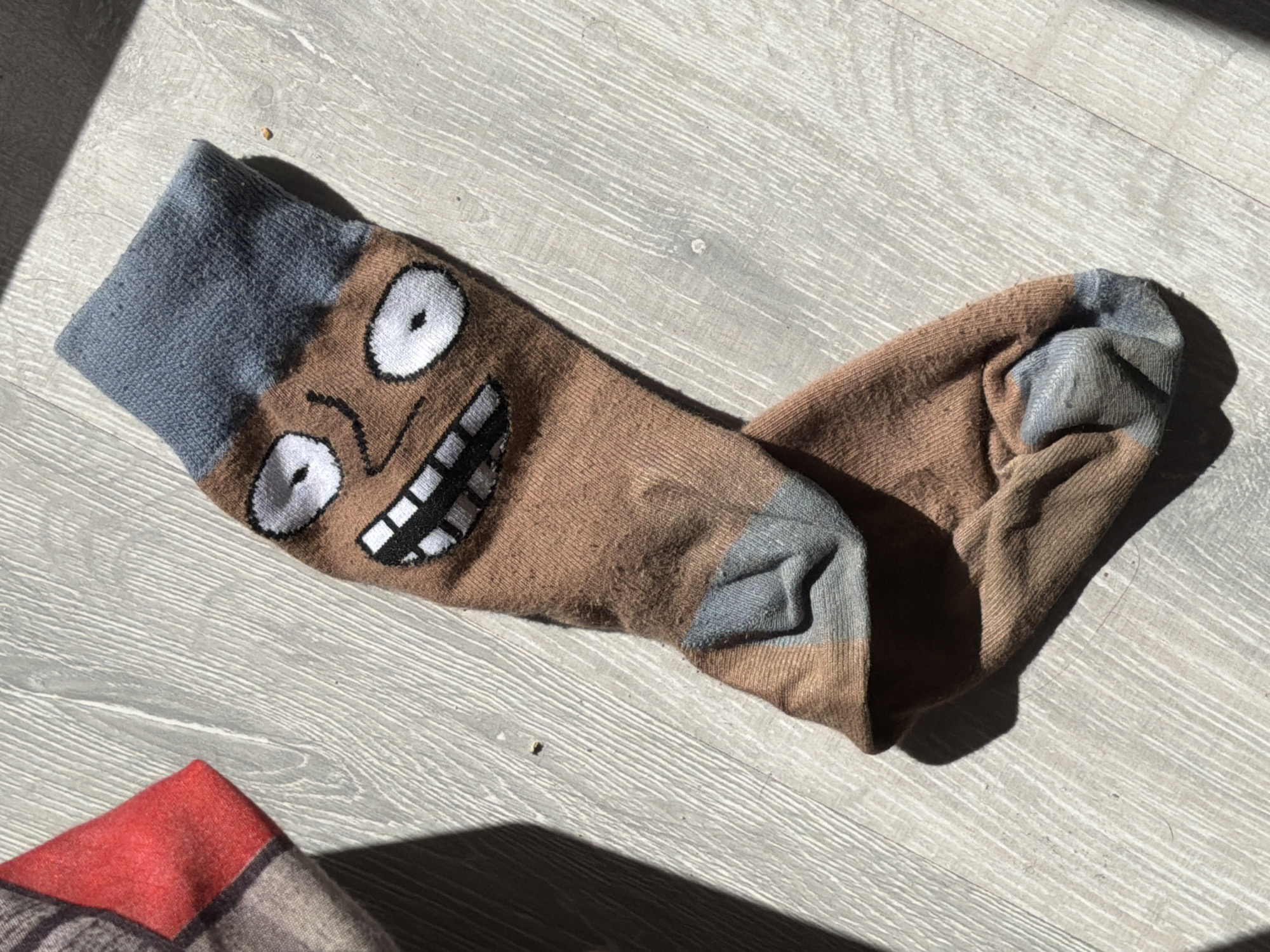 The image shows a brown sock with a cartoon face on a light grey wooden surface. The sock has a grey cuff and a brown body with a cartoon face featuring large white eyes, a simple nose, and a wide grin with visible teeth. Another object with red coloring is in the lower left corner of the image. The environment is well-lit, suggesting daylight. The exact location within Saint Petersburg, Russia, cannot be determined from the visual information alone.