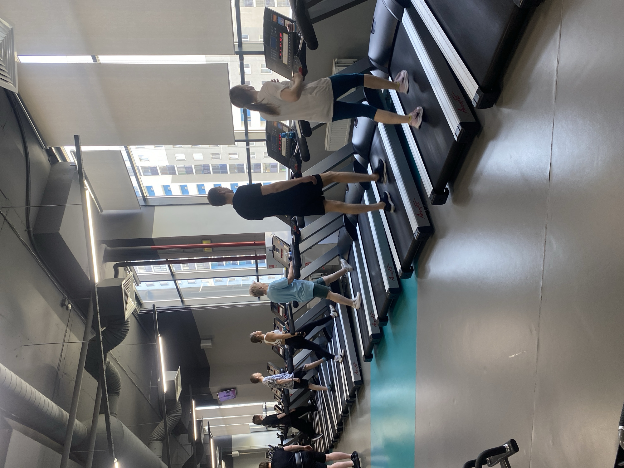 Several people are exercising on treadmills inside a gym. The treadmills are arranged in a row along a wall with windows. The windows provide a view of the city, which appears to be Yekaterinburg, Russia. The gym has high ceilings with exposed pipes and lighting fixtures. The walls are painted a dark gray color. The floor is a mix of gray and light teal. The people on the treadmills are wearing athletic clothing and running.