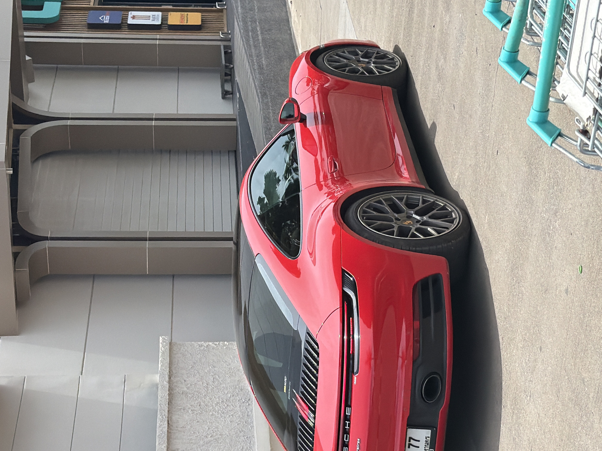 A red Porsche 911 Carrera is parked on a concrete surface in front of a modern building with large arched entrances. The car is facing away from the camera, showing its rear and right side. The building's facade is light-colored with horizontal paneling, and there are dark vertical elements to the right of the entrances, some of which appear to be signage. In the foreground, two turquoise shopping cart handles are visible, suggesting the car is parked near a shopping area. The lighting indicates it is daytime, with shadows cast on the ground. The scene appears to be in a commercial or public space within the city of Ko Samui, Thailand, based on the license plate which displays Thai script. There are no people visible in the image.