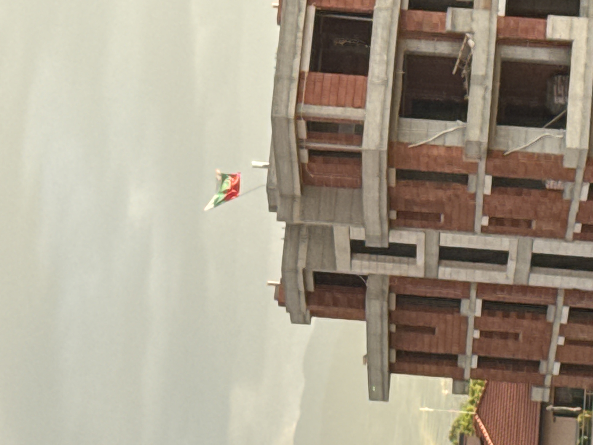 A tall building is under construction, with exposed concrete and red brickwork. The sky is overcast and pale. A small flag with green, red, and white colors flutters to the left of the building. The location is Unknown, Unknown.