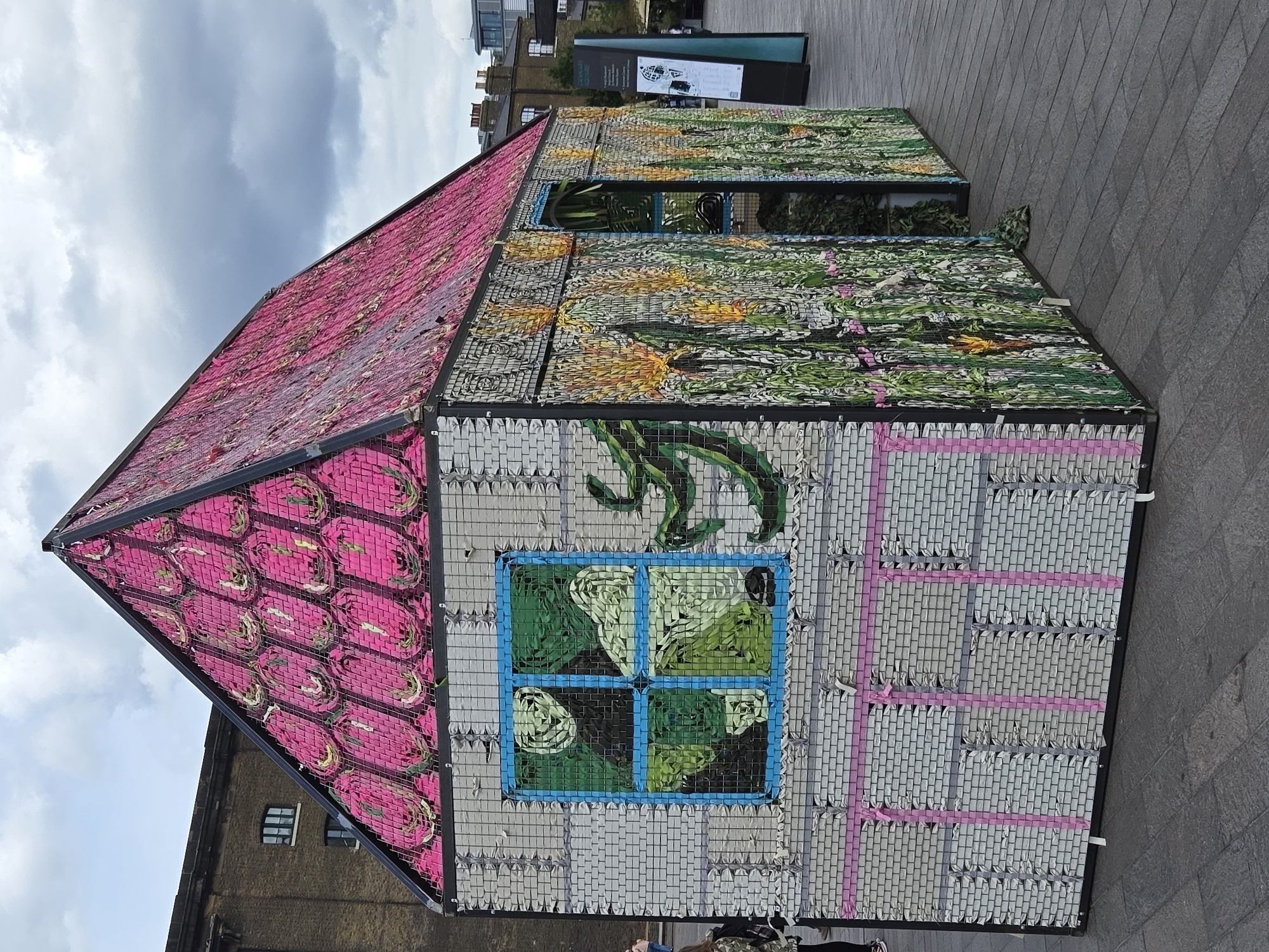 The image captures a distinctive art installation of a small house located in what appears to be a public space. The house is constructed from what seem to be upcycled materials, potentially plastic waste, arranged in a brick-like pattern. The roof is covered in pink material, while the sides feature a mix of colors and designs, including green foliage and a blue-framed window with abstract patterns. The environment is overcast, with a glimpse of buildings and informational signs in the background. The overall setting suggests the scene is an urban outdoor space in a city. The house is located near King's Cross, London.