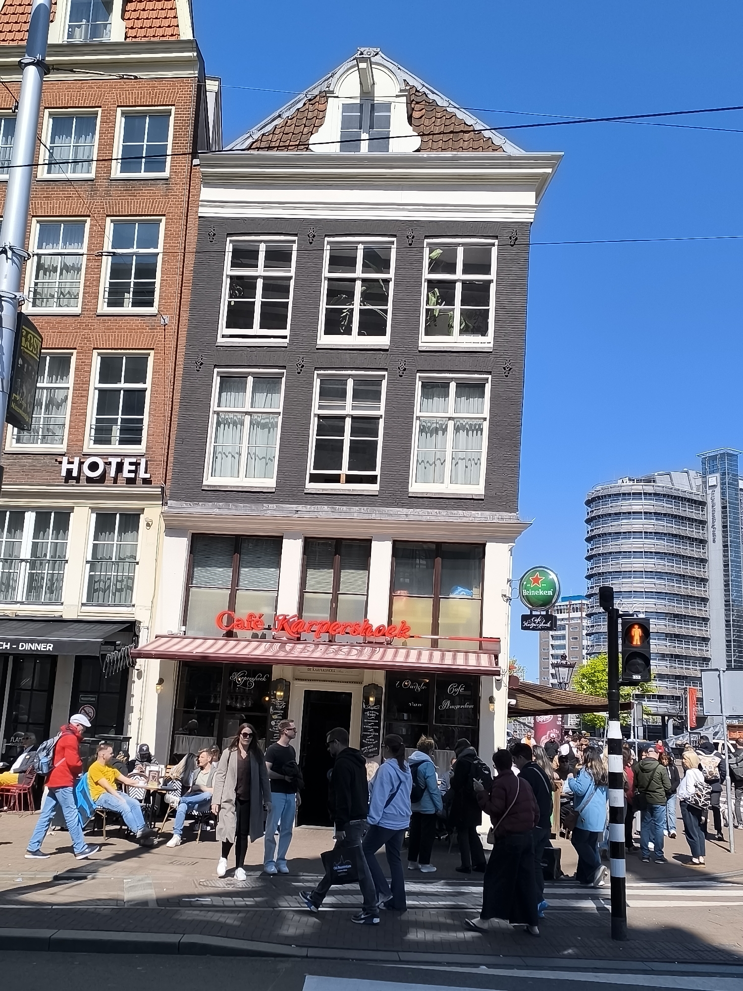 The image shows an urban street scene in Amsterdam, Netherlands, crowded with pedestrians crossing the street at a crosswalk. The building in the foreground is the "Cafe Karpershoek", with red and white awnings. To the left is a hotel, and a restaurant with black awning labeled "CH - DINNER". Behind the Cafe Karpershoek is a modern, multi-story building. The sky is clear blue. A traffic light shows a red signal. People are walking, sitting, or standing, engaged in conversations or waiting to cross.