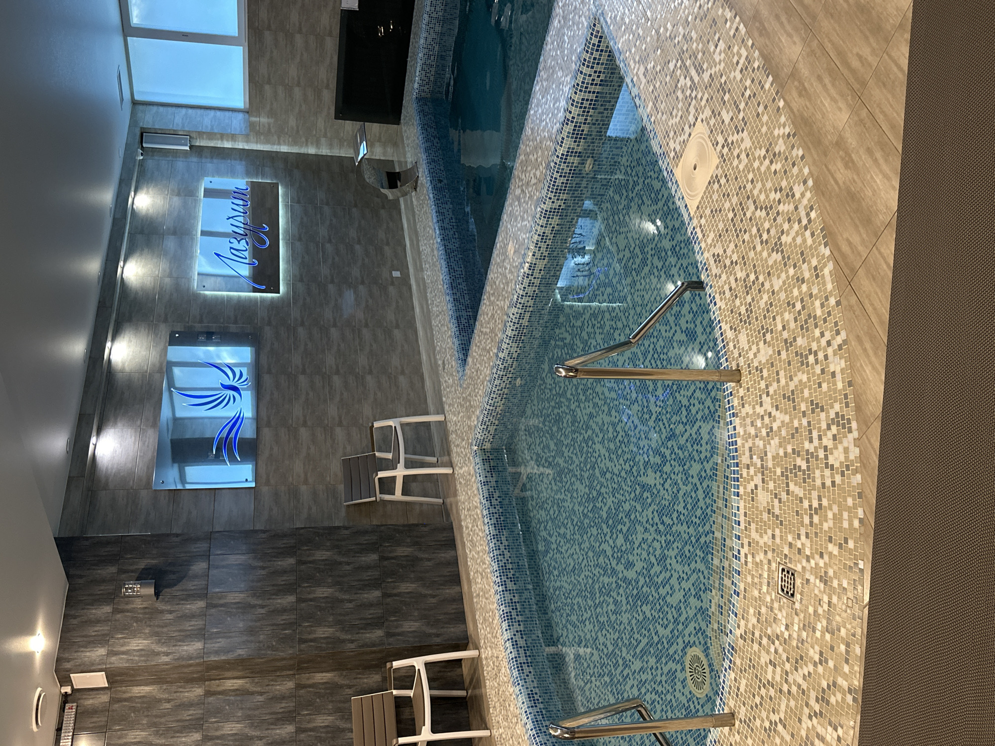 The image depicts an indoor spa area with two pools. The foreground features a smaller, shallow pool with blue tiled walls and a mosaic tile border. A metal railing is visible on the left side of this pool, with a drain grate on the floor. Adjacent to this is a larger, deeper pool, also with blue tiled walls and a similar mosaic border. A modern, chrome water feature is present at the edge of this larger pool. The walls of the spa are clad in dark grey textured tiles. Two illuminated signs are mounted on the wall: one with a blue bird graphic and another with blue Cyrillic lettering that reads "Лазурит" (Lazurit). Two white chairs with grey seating are positioned on the tiled floor between the pools and the wall. The environment is well-lit with recessed ceiling lights. Large windows with opaque blue coverings are visible on the right side of the image.