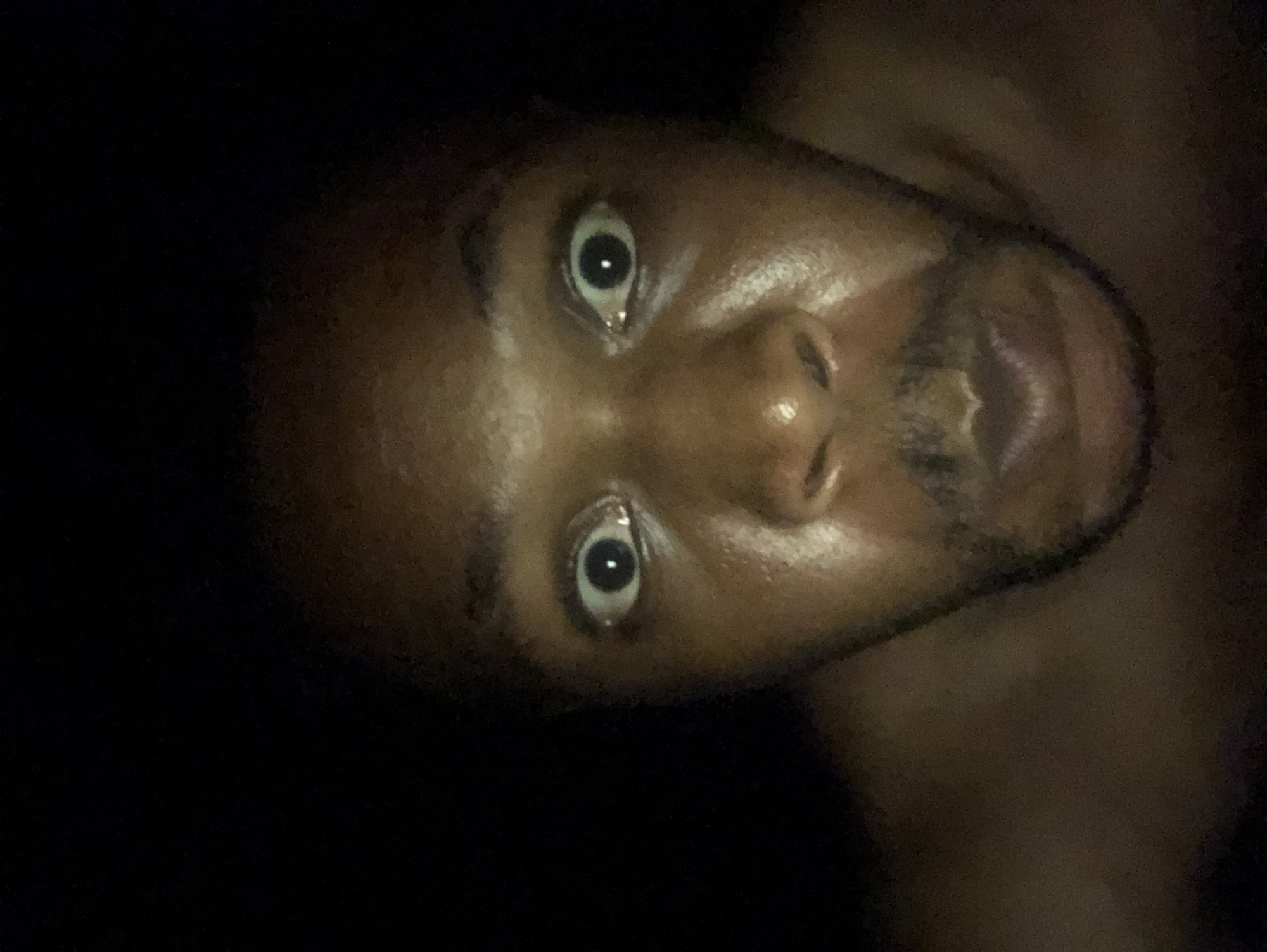 The image shows a close-up of a person's face in low light. The person has dark skin, wide eyes, and a visible beard. The background is dark and indistinct.