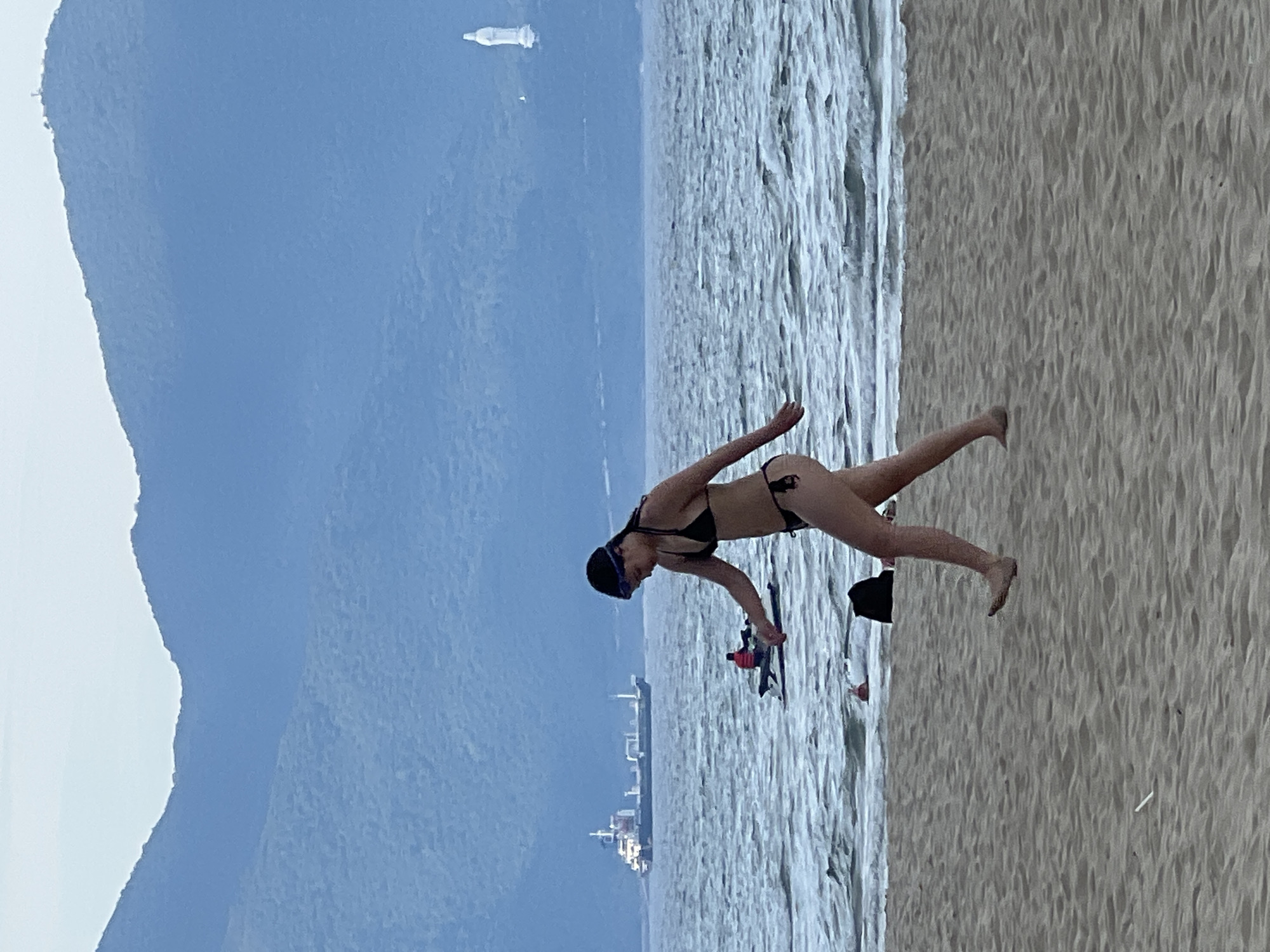 The image captures a scene at a beach in Da Nang, Vietnam, with a prominent mountain range in the background. In the foreground, a young woman wearing a black bikini and blue swimming goggles walks along the sandy shore. She is depicted mid-stride, with her body angled towards the right of the frame. The sea is visible behind her, with moderate waves and a few small boats in the distance. A large cargo ship is also visible on the horizon. Further back, a hazy white statue stands atop a distant hill. The sky is overcast, casting a soft light over the scene. The sand in the foreground shows textures of dunes and small debris.