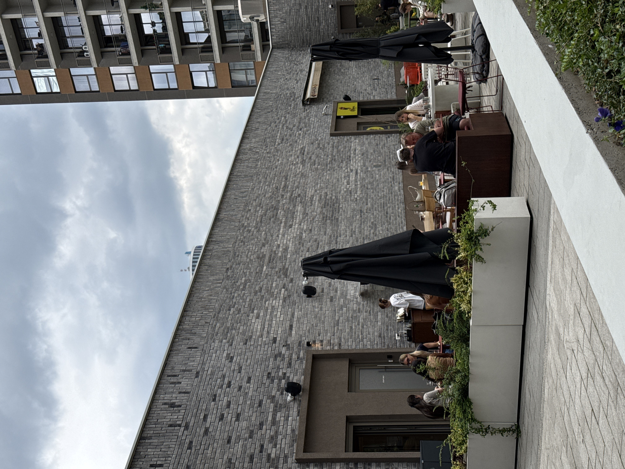 The scene depicts an outdoor seating area of a restaurant, likely in Belgrade, Serbia, given the architectural style. Diners are seated at tables under large black umbrellas. The restaurant features a grey brick exterior. A tall building is visible in the background under a cloudy sky. The tables are near a white planter. The foreground includes a stone-paved walkway.