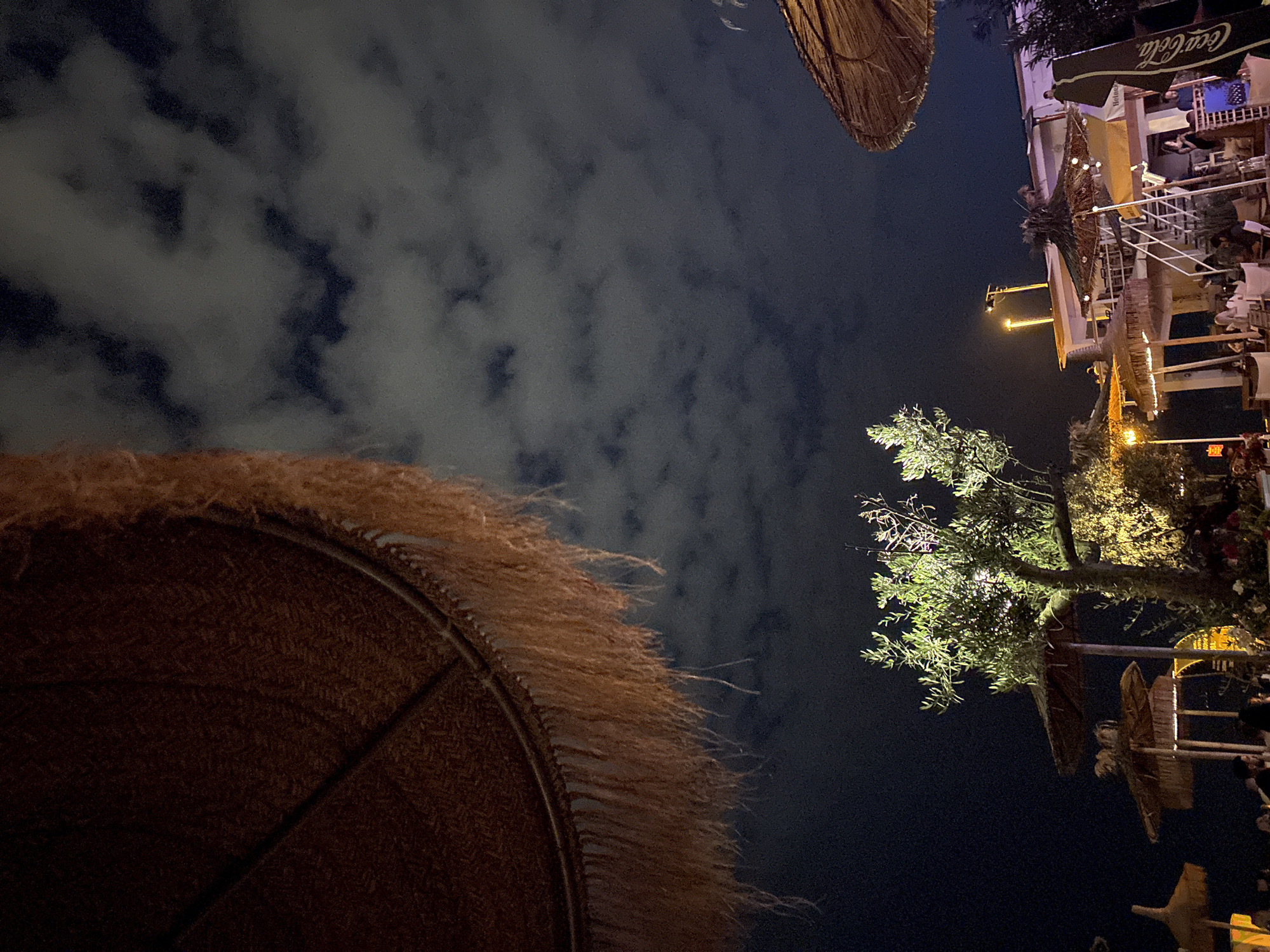 An outdoor scene at night, likely a beachside establishment. The foreground is dominated by the underside of a large, thatched umbrella with a fringed edge, positioned to the left and top of the frame. In the middle ground, a small, illuminated tree with lush foliage stands prominently. Behind the tree, several more thatched umbrellas and structures are visible, along with a white building with outdoor seating and a staircase. People are scattered throughout the scene, seated at tables and walking around. A tall, black Coca-Cola branded umbrella is visible on the right side of the frame. The sky above is dark with scattered clouds, illuminated by what appears to be moonlight or artificial lighting from the establishment. The overall impression is that of a lively, nighttime gathering at a resort or beach bar.