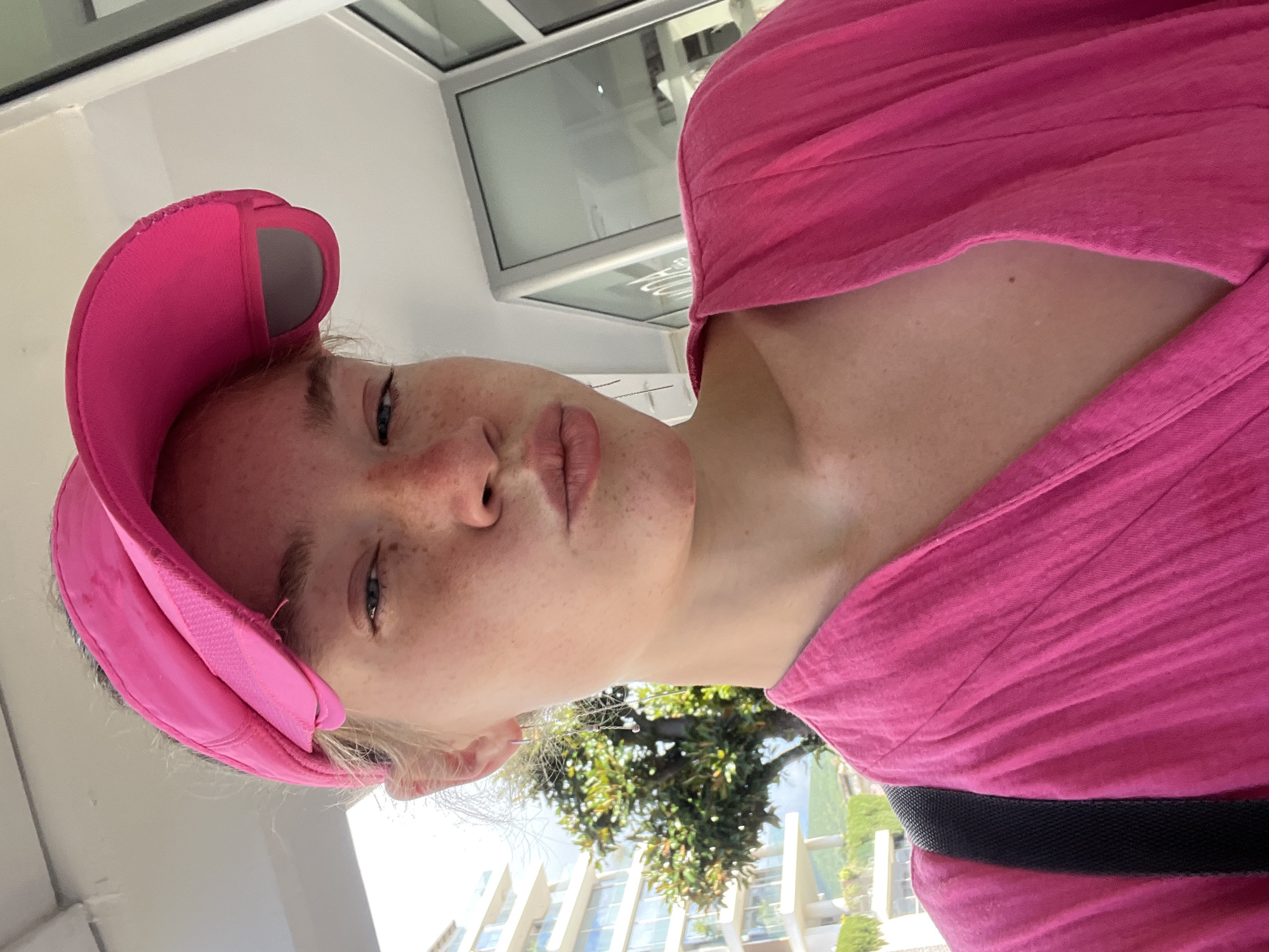 A young person with fair skin and light-colored hair, wearing a bright pink fabric visor and a matching pink top, looks directly at the camera. The subject has visible freckles across her nose and cheeks, and her lips are slightly parted. The image is taken from a low angle, with the person positioned in the foreground.
The background reveals an outdoor setting on a sunny day. Above and behind the subject, modern buildings with multiple large, rectangular windows are visible, reflecting bright light. Green foliage from trees or shrubs is visible in the middle ground, indicating a lush environment. A dark strap, possibly from a bag, crosses the subject's right shoulder and chest area. No specific activity is depicted other than posing for the photograph. There is no visible text in the image. The context suggests the location is Budva, Montenegro.