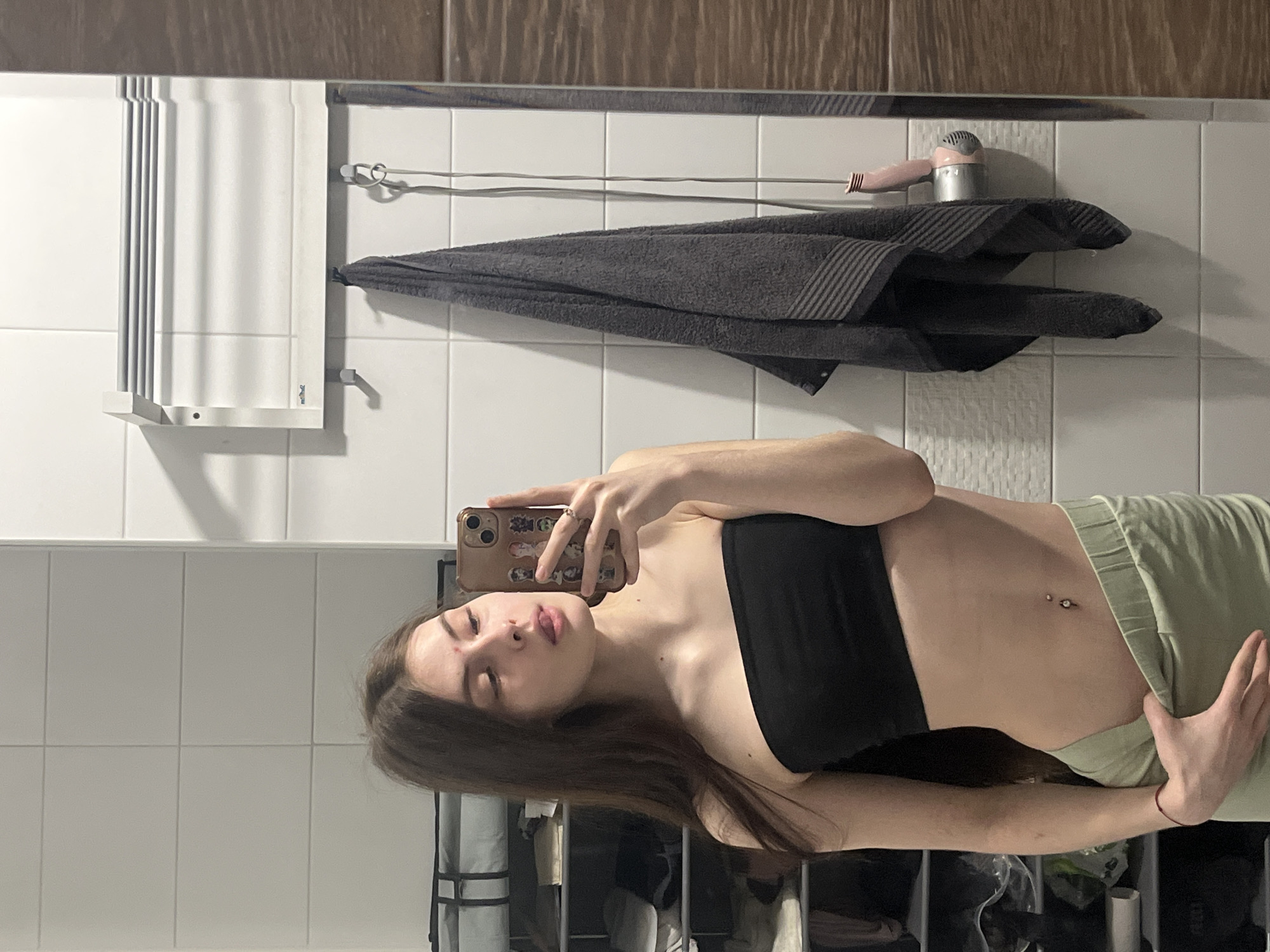 A young woman is taking a selfie in a bathroom. She is wearing a black tube top and light green sweatpants. Her phone has a decorative case. In the background, there is a tiled wall, a towel rack, a gray towel, and a hair dryer. A storage unit containing various items is visible beneath the woman. There is no indication of Saint Petersburg, Russia.