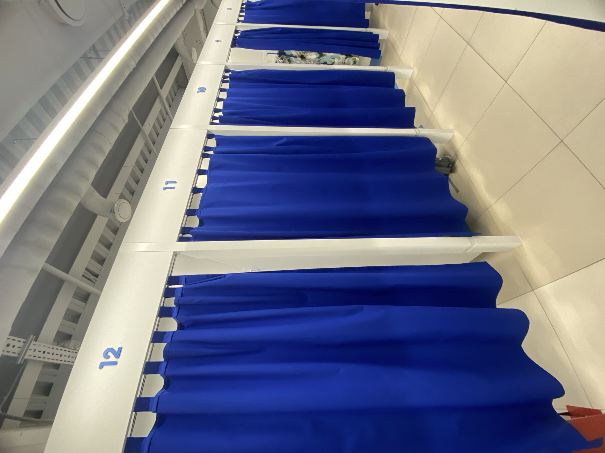 The image shows a row of numbered, curtained cubicles in Sochi, Russia. The cubicles are numbered 8 through 12, with blue curtains hanging from each. The cubicles are white, and the walls are tiled. The scene is well-lit.