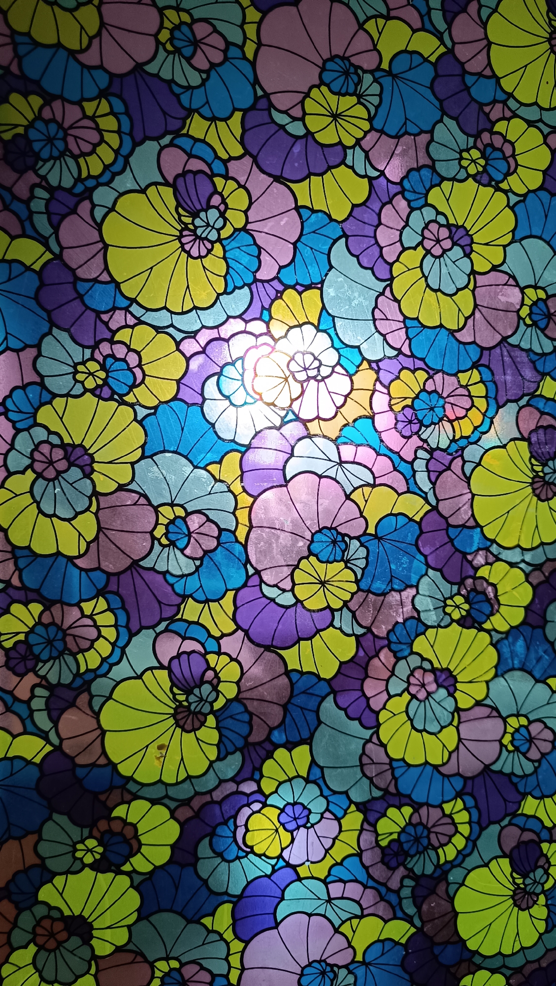 The image displays a stained glass window with a floral pattern. The flowers are stylized and overlap each other, creating a dense, mosaic-like effect. The colors are vibrant and include shades of purple, blue, yellow, pink, and green. The glass is illuminated from behind, causing some areas to be brighter than others, with a strong light source visible in the upper center of the image. There are no people, specific objects, actions, interactions, or environmental details that would indicate a location within the city of Karsha, Russia. The image is solely a close-up view of the stained glass artwork.
