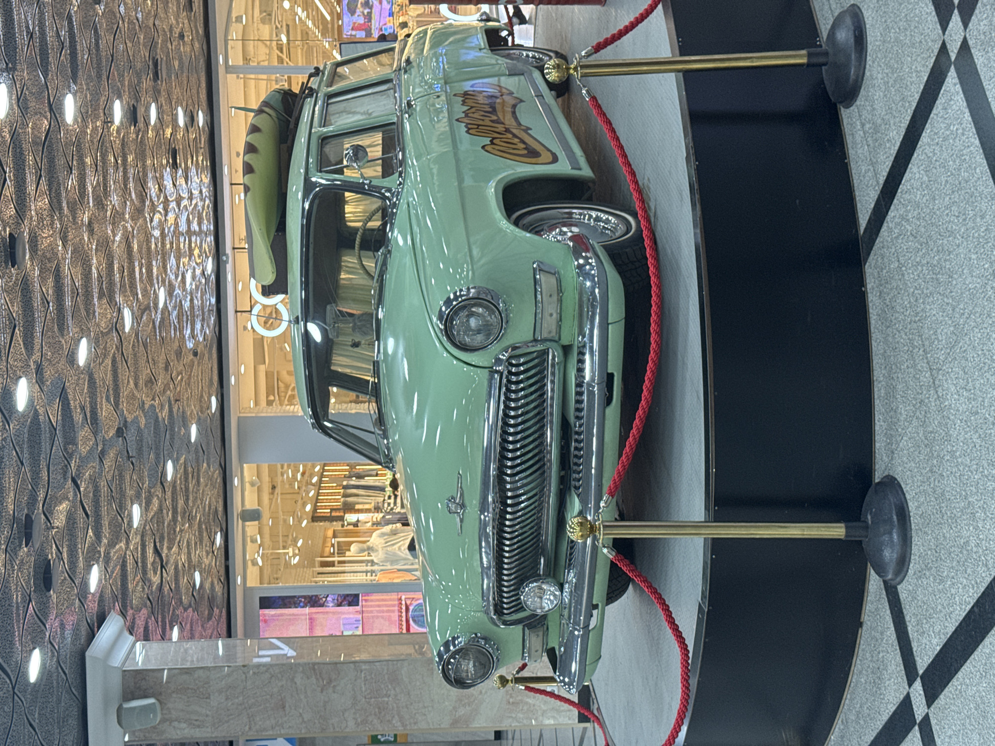 The image shows a vintage, light green station wagon with a surfboard on its roof, displayed inside a shopping mall in Yekaterinburg, Russia. The car is positioned on a raised black platform, surrounded by red ropes held up by golden stanchions. The car has chrome detailing on the grille, headlights, and wheels, and the word "California" is written in yellow and brown lettering on its side. The mall environment is visible in the background, with shops and mannequins. The ceiling of the mall features an intricate design with embedded lights. The floor is tiled with a checkered pattern. The overall scene depicts a car exhibition or display within a commercial setting.