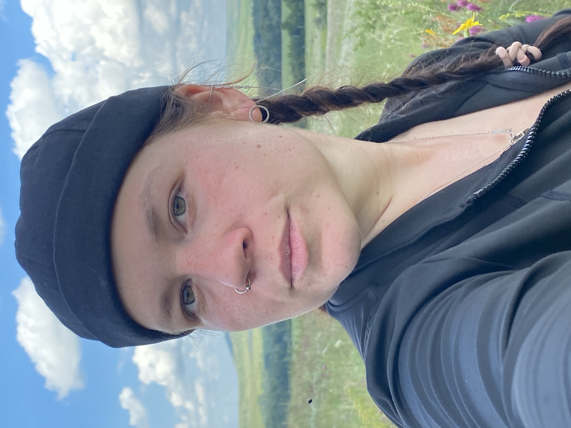 The image shows a close-up, selfie-style shot of a woman with light skin, green eyes, and light brown hair styled in a braid and worn under a black beanie. She has a nose ring and earrings. She is wearing a black zip-up jacket. The background is a landscape with green hills, scattered trees, and a blue sky with white clouds, suggesting an outdoor setting, possibly in or near the village of Ziyanchurino, Russia, given the open, rolling terrain. The focus is on the woman's face and upper body, with the landscape softly blurred.
