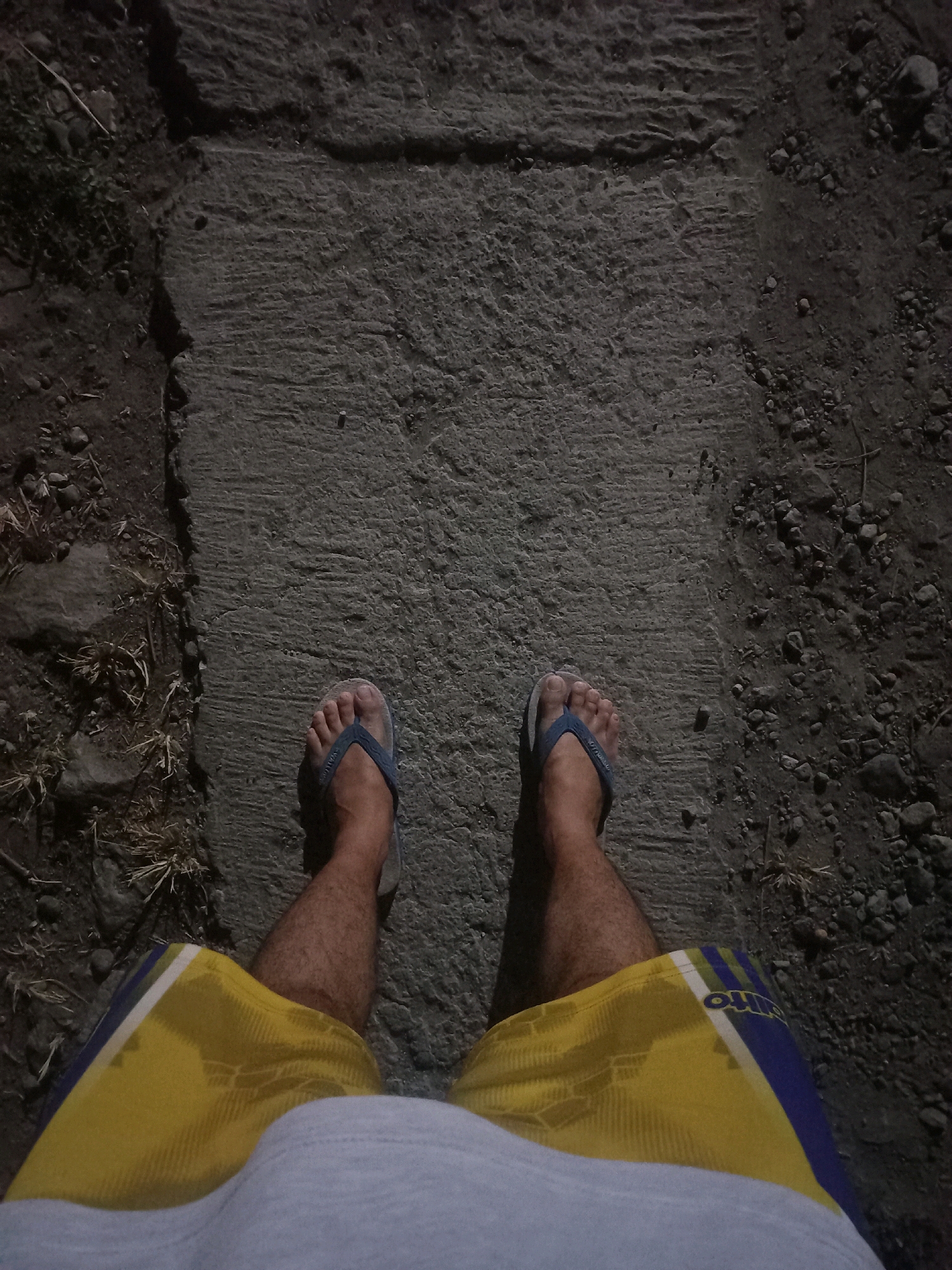 The image shows a first-person perspective of someone standing on a concrete pathway in Gambiranom, Indonesia. The pathway is cracked and bordered by rough dirt and small stones. The person is wearing blue flip-flops and yellow shorts with blue and white stripes. The lighting suggests it is nighttime or dusk.