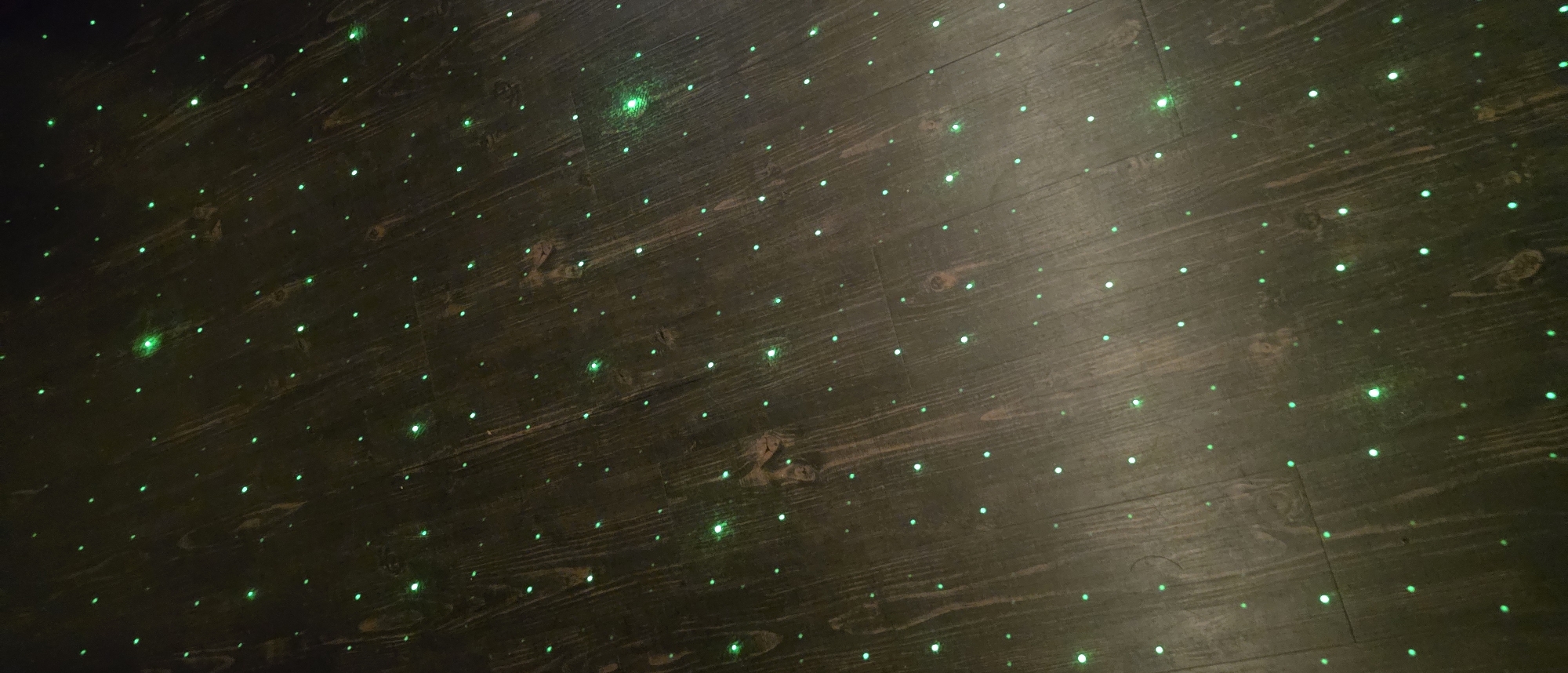 The image shows a dark, wooden floor covered in evenly spaced, small, green dots of light. The floor appears to be made of planks and has a slightly glossy finish. The scene is dimly lit, with the green lights providing the primary illumination. The image does not provide information about the location in Sapareva Banya, Bulgaria, or about people, specific subjects, objects, actions, or interactions.