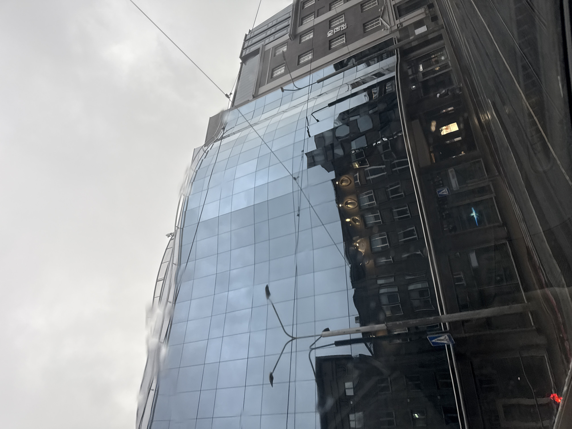 This image shows a view of the city of Tallinn, Estonia, captured from within a vehicle, likely a tram. The primary subject is a modern glass-fronted building that reflects the surrounding environment, including another, older building. The sky is overcast and gray, suggesting a cloudy or rainy day.
The modern building is characterized by its large, reflective glass panels that create distorted reflections of the street, a street lamp, and the adjacent building. The older building, to the right of the modern one, is made of stone or brick and has a more traditional architectural style with multiple windows and decorative elements.
In the reflection of the modern building, the opposite side of the street is visible. This includes the facade of another building with windows and what appears to be some lighting or signage. A street lamp, with two lamps on either side of a central pole, is reflected prominently in the glass.
On the street level, tram tracks are visible, indicating that the photograph was taken from a tram or a similar vehicle. The street appears wet, which could be due to rain or recent cleaning, contributing to the reflections. A blue pedestrian crossing sign is visible on the side of the road.
In the foreground, the interior of the vehicle from which the photo is taken is partially visible, suggesting a window view. The overall impression is of an urban street scene in a European city, with a blend of modern and older architecture. The lighting is diffused and somewhat dim due to the overcast weather.