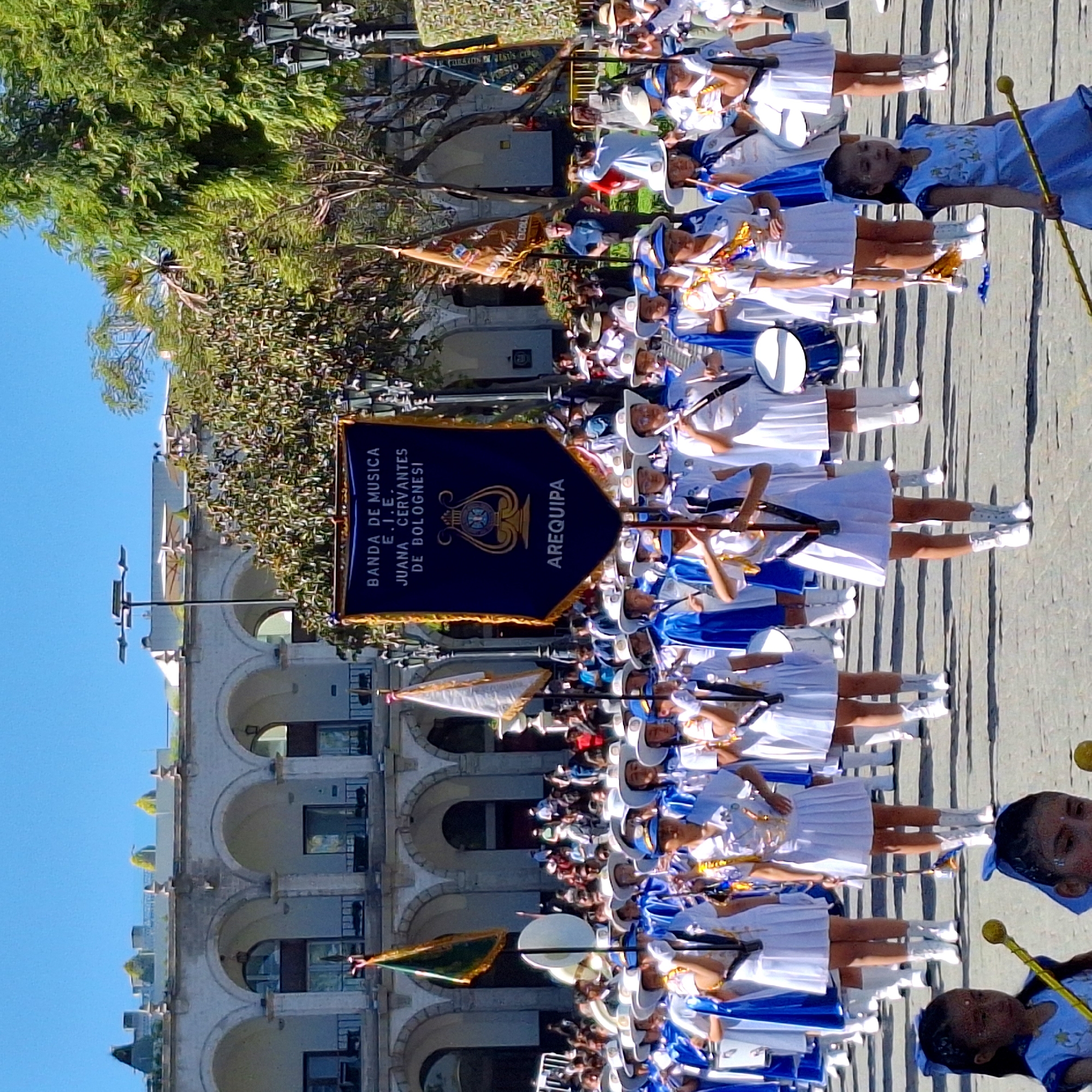 Captured from a ground-level vantage point, the image showcases a marching band, clad in white and blue uniforms, parading along a city street. The band members, positioned in neat rows, are playing their instruments, some holding flags or batons. A prominent navy blue banner, emblazoned with the words "BANDA DE MUSICA E.I.E. JUANA CERVANTES DE BOLOGNESI" and "AREQUIPA," is held high. The backdrop features a stone building with arched windows, a tree with dense foliage, and a clear blue sky. The scene is set in the city of Arequipa.