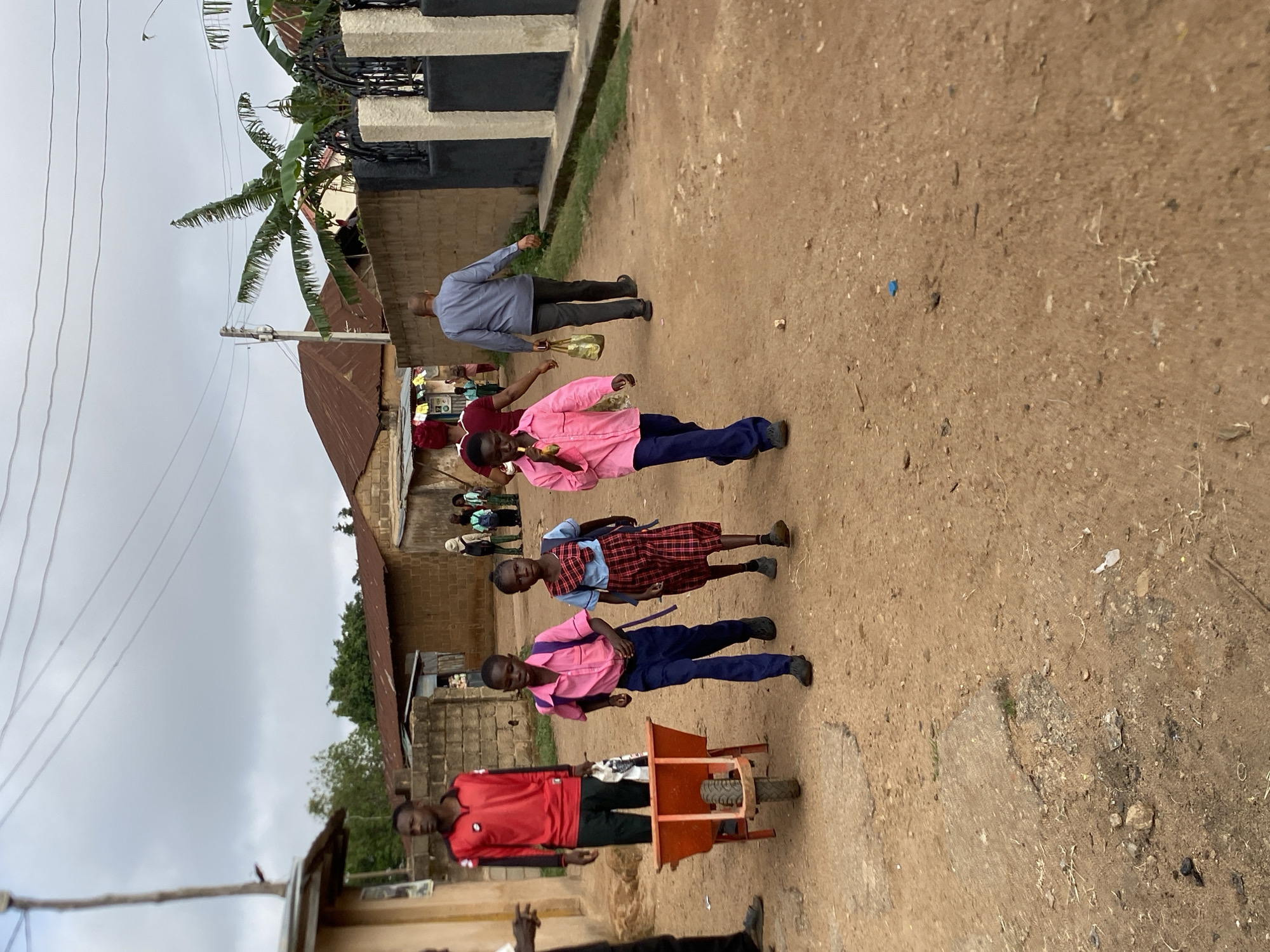 In Bukuru, Nigeria, a street scene depicts several individuals walking along a dirt road. At the front, a person in a red shirt pulls a small, orange wheeled cart. Behind them are two children in school uniforms: the first in a pink shirt and blue pants, and the second in a red plaid dress with a blue shirt. Further back, another person in a pink shirt and blue pants walks along the road, with an additional person in a blue shirt visible in the background. Houses and buildings with corrugated iron roofs line the road, and a tall palm tree rises behind one of the buildings. Overhead, the sky is overcast with light gray clouds.