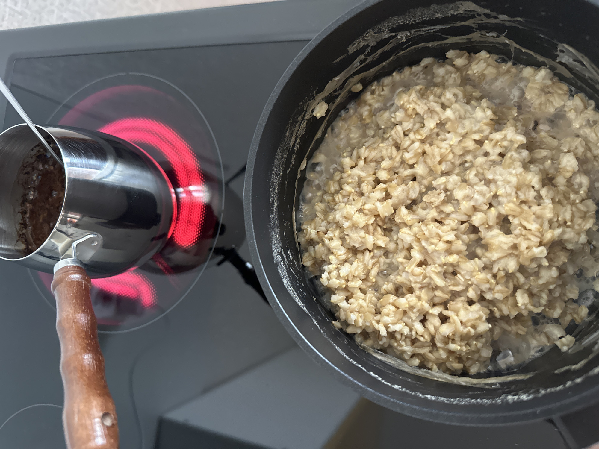 The image depicts a close-up, high-angle view of a stovetop with two cooking vessels. In the foreground, a black pot filled with cooked oatmeal is shown, with some of the oatmeal having spilled over the rim. In the background, a small silver pot with a wooden handle, containing a dark liquid, sits on a glowing red electric burner. The scene appears to be in a kitchen environment, but its specific location within the city of Yuzhnouralsk, Russia, cannot be determined from the visual information provided. There are no people visible in the image.