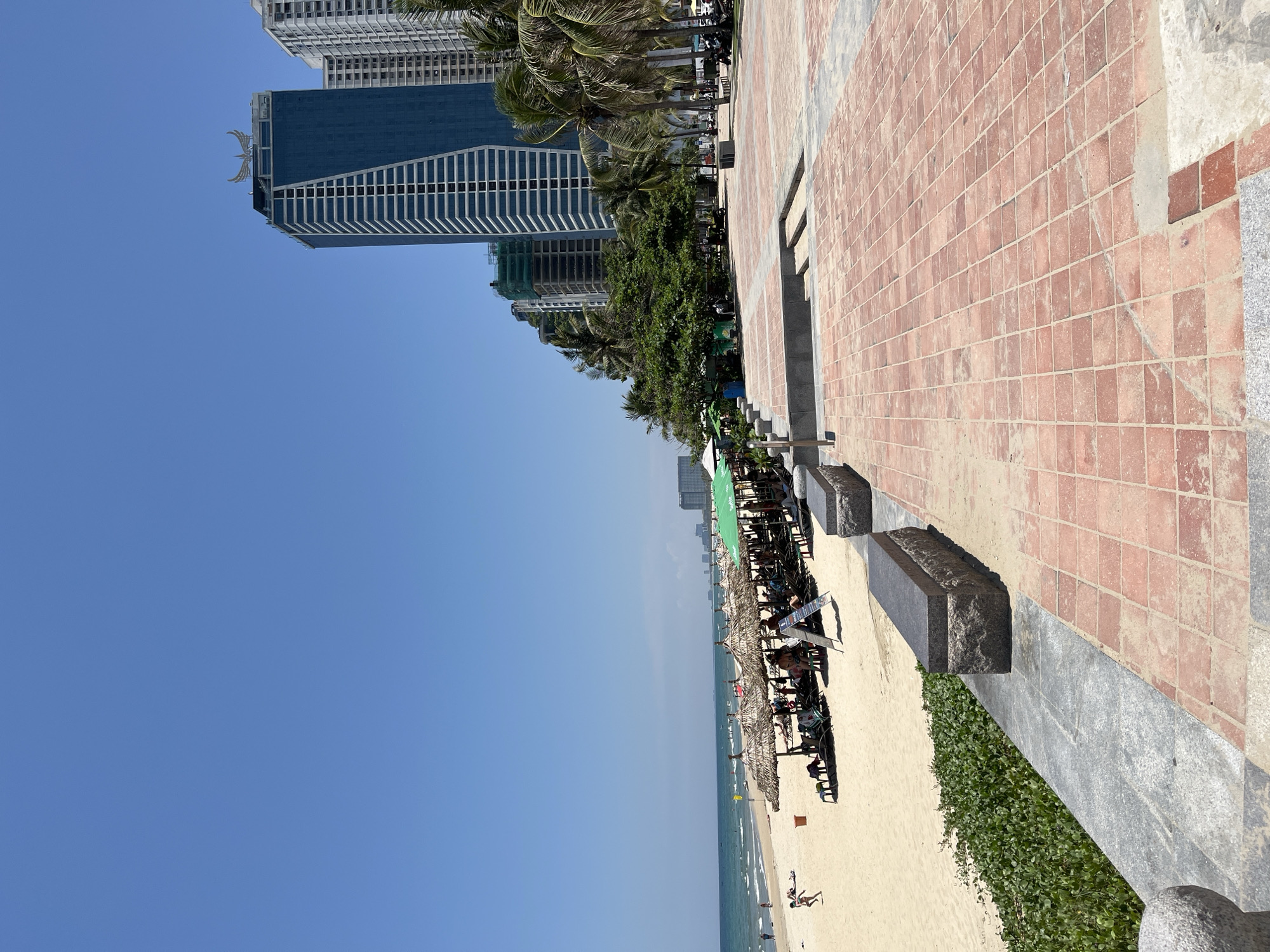 The image depicts a coastal scene in Đà Nẵng, Vietnam. On the right, a brick walkway transitions to a sandy beach with lounge chairs and thatched umbrellas. Palm trees and skyscrapers line the background. The blue sky occupies a significant portion of the frame. People are present on the beach, with some appearing to be swimming or sunbathing.