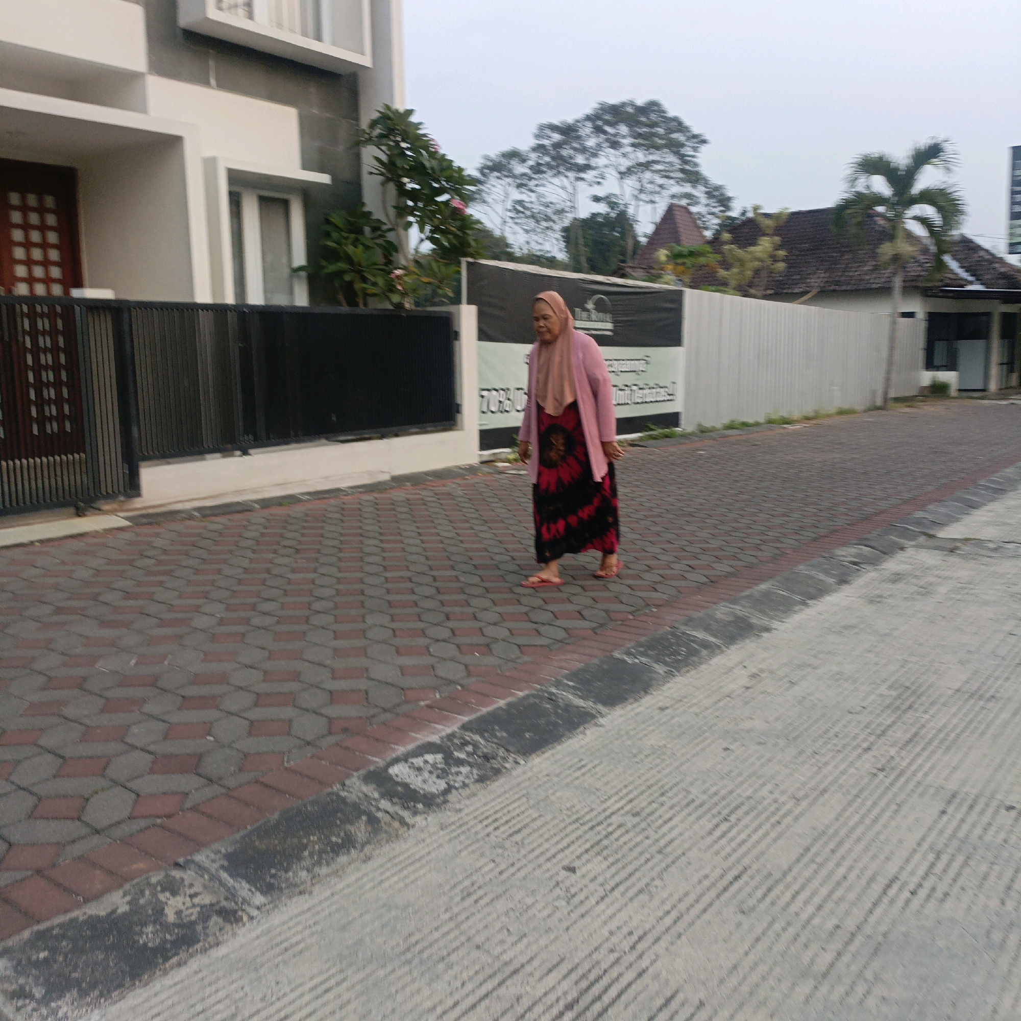 The image shows a woman walking on a patterned brick sidewalk in front of a building with a dark metal fence. The woman is wearing a pink hijab and a pink jacket over a black and red patterned dress, and she is wearing red flip-flops. Behind the fence is a modern building with white accents and a dark door. Further in the background, there is a sign with text, trees, and houses with tiled roofs. The sky is overcast. Based on the architectural style, vegetation, and the presence of a sign with Indonesian text, this image is likely from Salatiga, Indonesia.