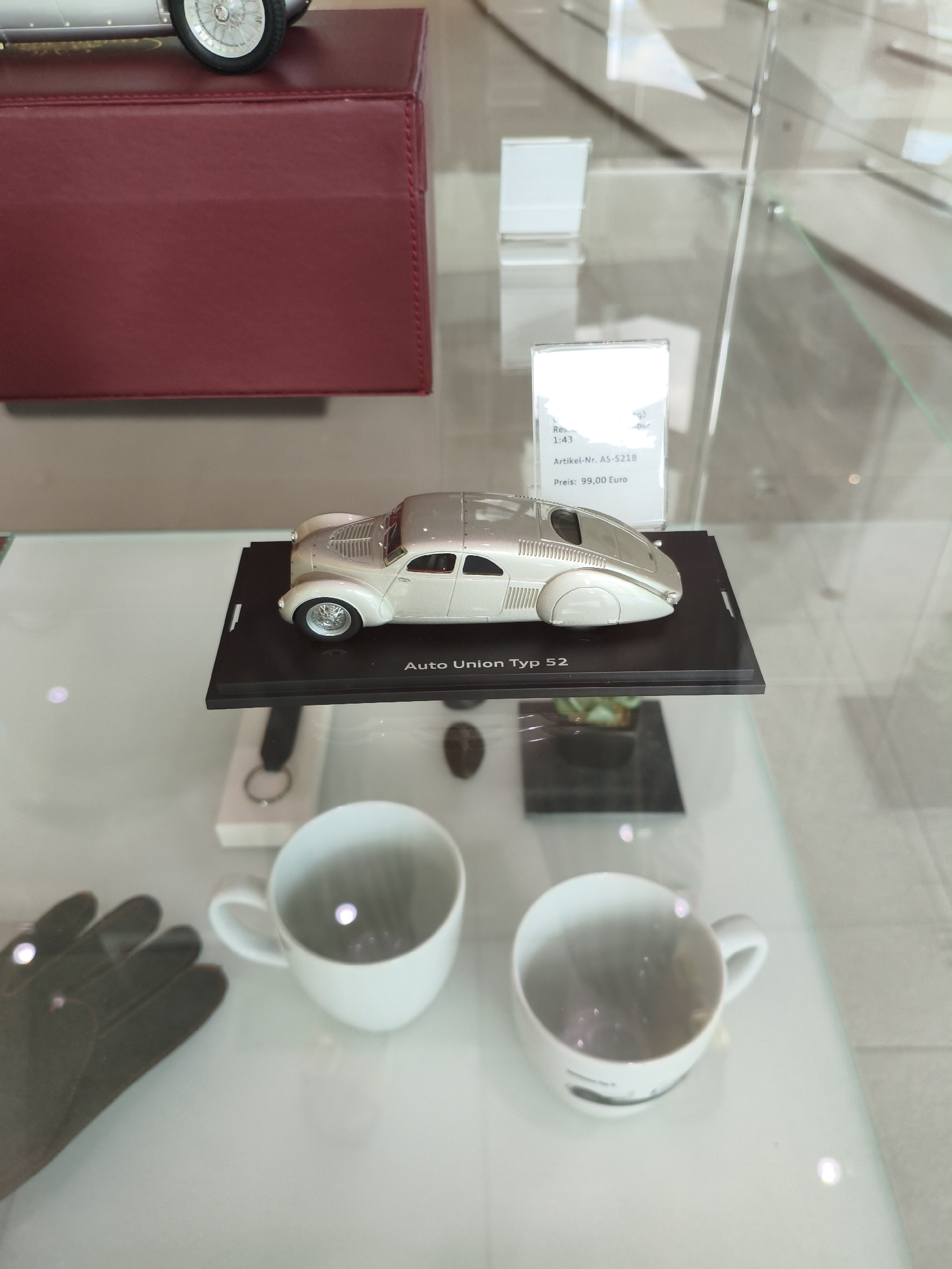 The image depicts a display case within a museum or showroom, likely located in Ingolstadt, Germany, given the presence of an Auto Union branded item. The display case is made of glass and shows a model car, specifically an "Auto Union Typ 52," on a black base. The model car is silver and appears to be in a scale of 1:43. Next to the model car, a label provides product details, including an article number "AS-5218" and a price of "99,00 Euro."
In the foreground, two white ceramic mugs are visible, one on the left and one on the right. A dark brown glove lies to the left of the mugs. The background shows reflections of the museum or showroom environment, including other display elements and possibly lighting fixtures. The overall lighting is bright, with reflections from the glass surfaces. The scene is static and presents a collection of items for viewing or purchase.