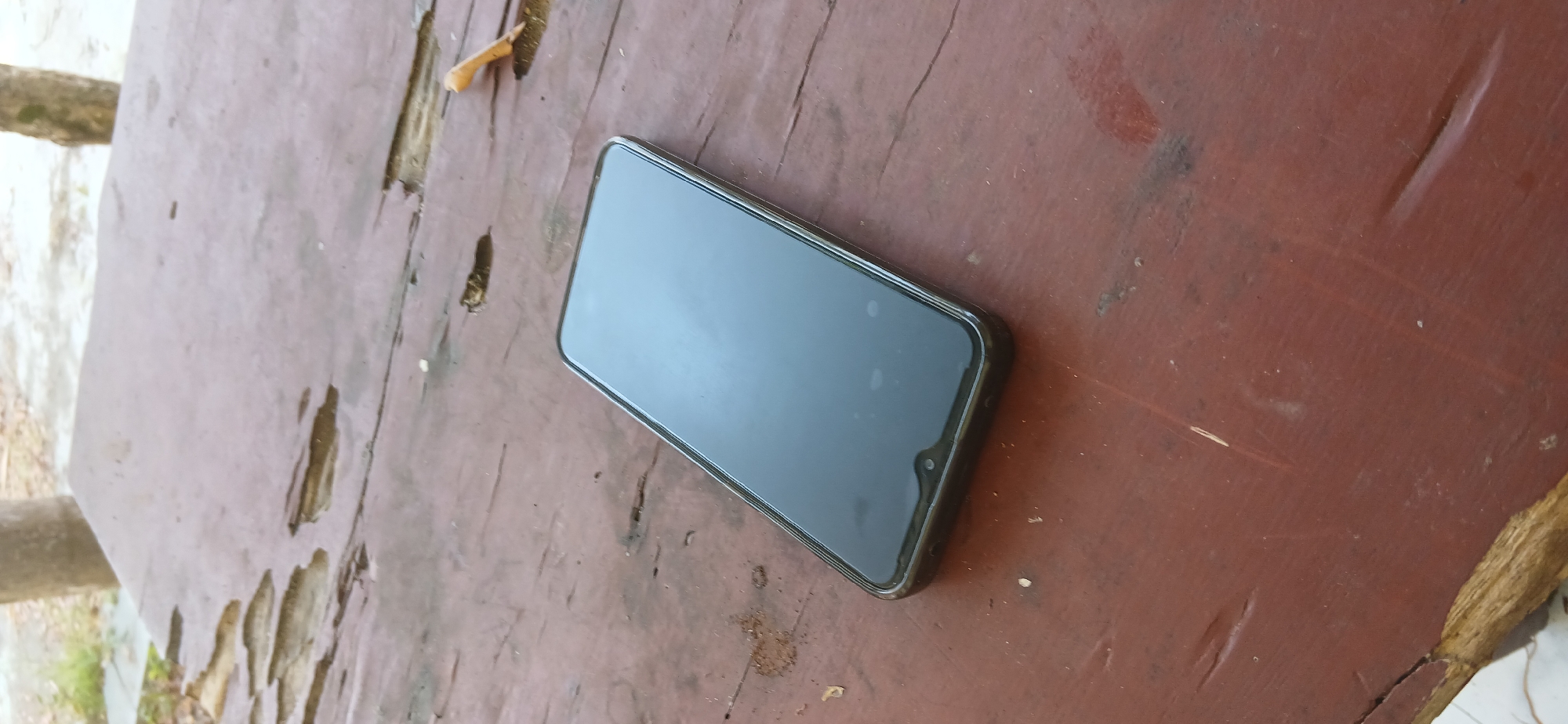 A dark-colored smartphone is resting on a dark-brown wooden table with a distressed, weathered finish. The table surface shows signs of damage, including chipped paint and worn areas, with visible wood grain underneath. In the background, the blurry figures of tree trunks are visible, suggesting an outdoor setting. A small, light-colored twig or piece of debris lies on the table to the right of the phone. The lighting suggests it is daytime, with soft light illuminating the scene. The smartphone's screen is dark and reflective, obscuring any content. The image does not contain any people, actions, or interactions. The location is specified as Pejaten, Indonesia.