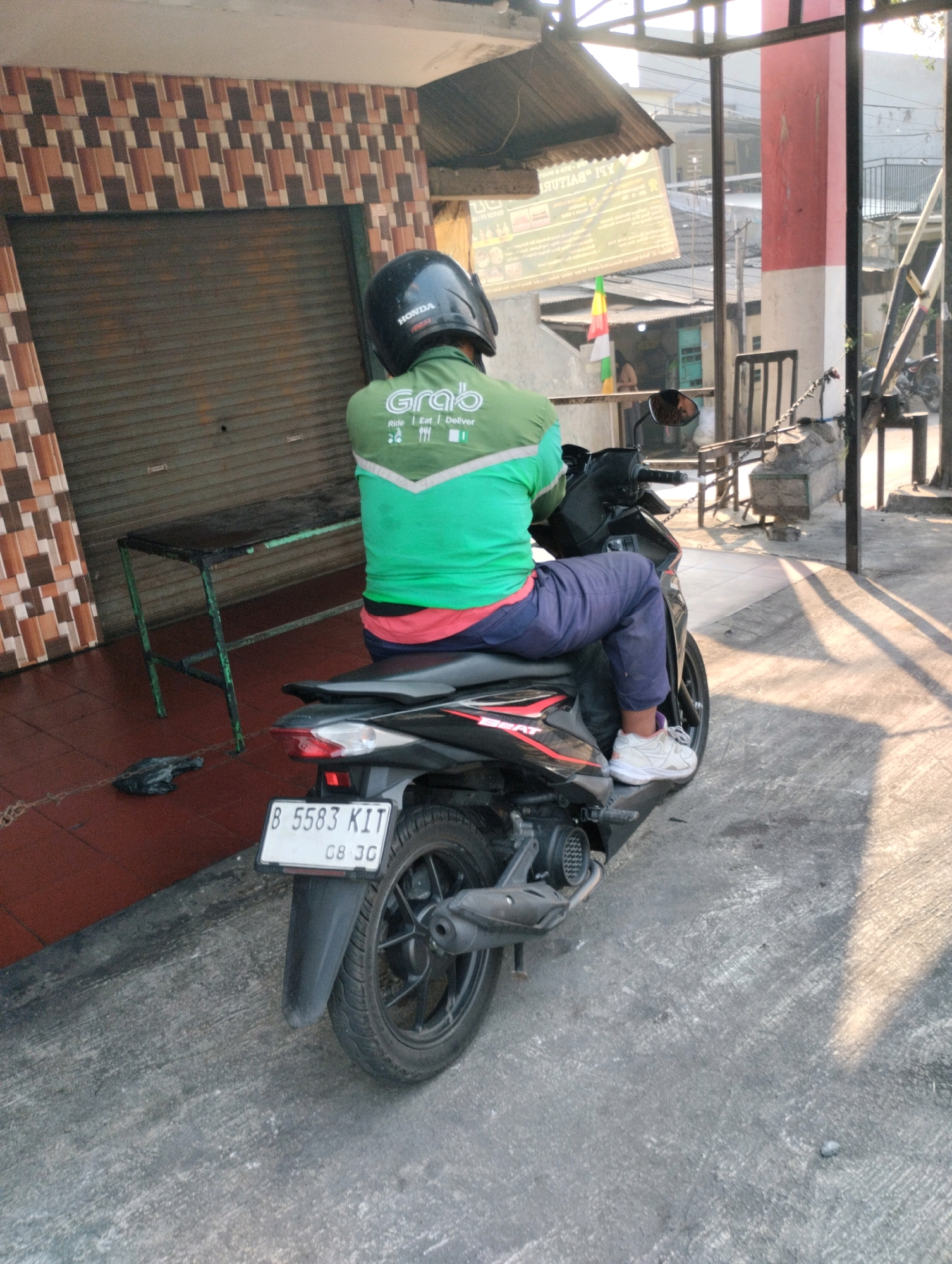 A person wearing a black helmet and a green Grab delivery uniform is sitting on a black motorcycle. The uniform has the Grab logo and the words "Ride | Eat | Deliver". The person is wearing blue pants and white sneakers. The motorcycle has a license plate that reads "B 5583 KIT" and "08.30". The scene appears to be an outdoor area in Bekasi, Indonesia, with a building with a patterned facade and a metal roll-up door in the background. A metal table is placed on the ground to the left of the motorcycle. The ground is paved with concrete and tiles. Sunlight casts shadows on the ground.