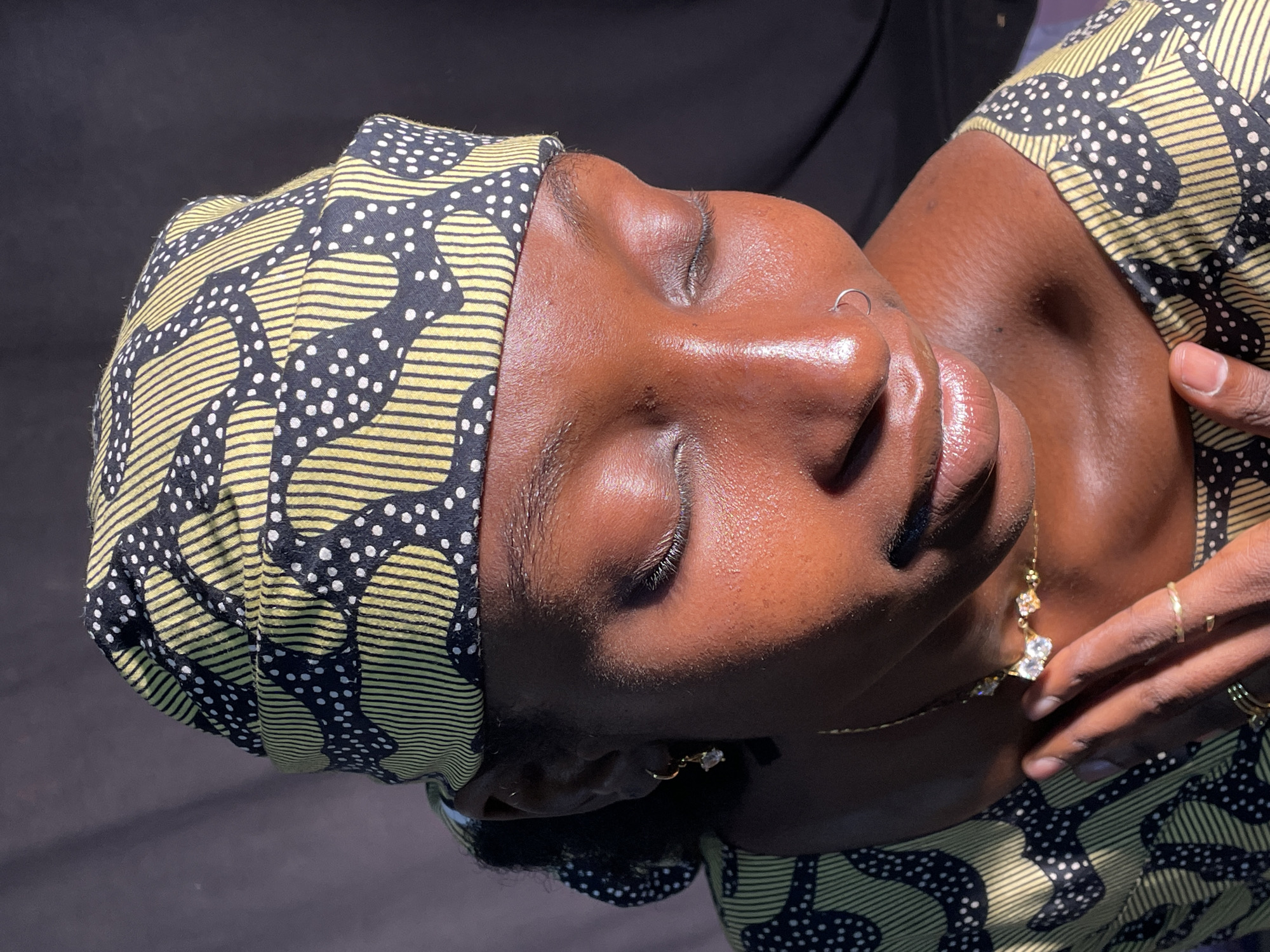 A close-up portrait shot features a woman with her eyes closed, illuminated by strong, directional light, creating highlights on her skin and casting shadows. Her head is tilted to the right, and her face is in profile or slightly angled. She has a smooth, dark complexion, well-defined eyebrows, and full lips. A small silver hoop nose ring adorns her left nostril.
She is wearing a vibrant headwrap and garment, likely made from the same fabric, which features a distinctive pattern of horizontal yellow stripes intertwined with black abstract shapes dotted with white. Gold-toned jewelry is visible, including small earrings with clear stones, a delicate necklace with a clear gemstone pendant, and multiple thin rings on her fingers. One hand, adorned with rings, is gently positioned near her shoulder or neck, suggesting a contemplative or relaxed pose. The background is dark and nondescript, focusing attention on the subject. There is no visible text. The overall scene conveys a sense of calm and serenity, possibly taken indoors with sunlight streaming in. The context provided indicates the location is Jos, Nigeria.