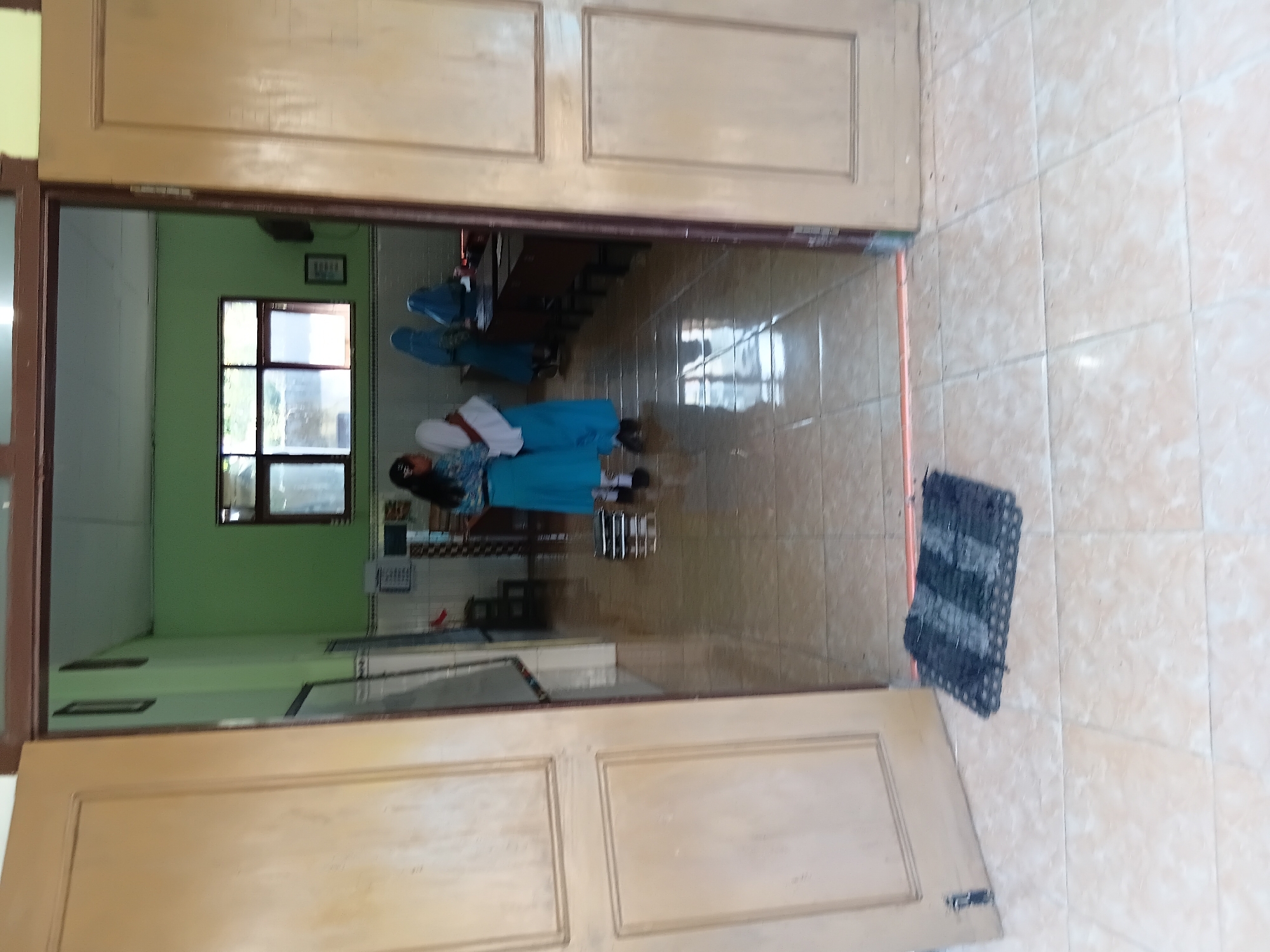 The image depicts an interior view of what appears to be a classroom or school hallway in Tanggul, Indonesia. Two young female students, dressed in blue and white uniforms, are standing in the middle of a hallway. One student is facing away from the camera, while the other faces partially towards it. Behind them, two more students, also in uniforms that include headscarves, are seated at desks in what looks like a classroom. The walls of the classroom are painted green, and a large window is visible, offering a view of an outdoor area. In the foreground, a doorway is open, revealing a tiled floor with a dark doormat. The overall environment suggests a typical school setting within the city of Tanggul.